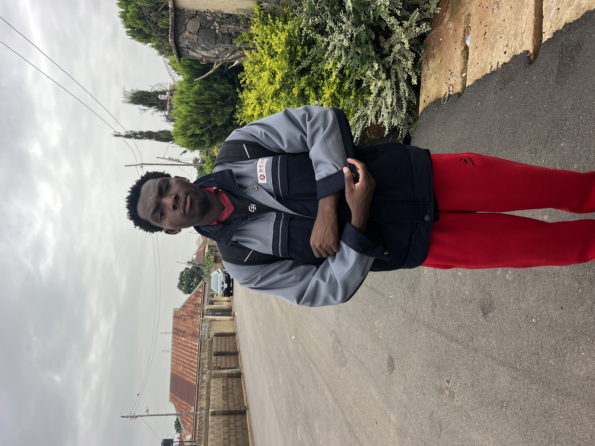 A young Black man stands on an asphalt road in front of a lush green bush and a stone wall. He is wearing a grey and navy blue jacket over a red shirt, and bright red pants. The man is looking directly at the camera with a neutral expression. Behind him, a road stretches into the distance, lined with houses and a fence. The sky is overcast. A black car is parked on the side of the road.