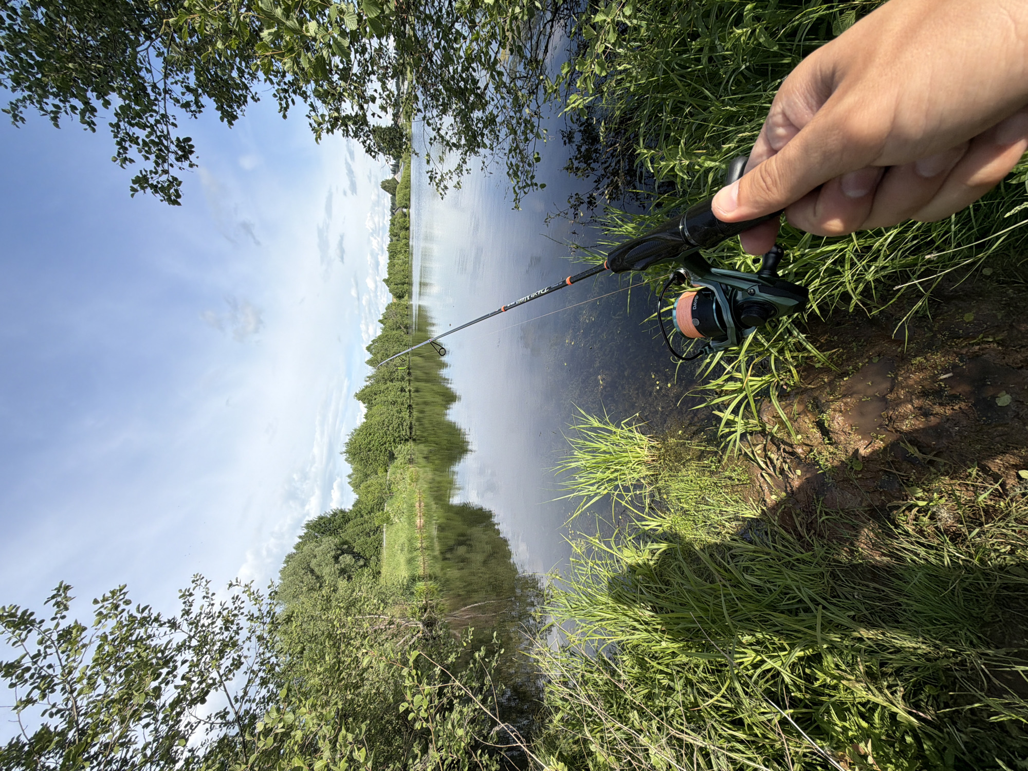A vertical-format outdoor photograph captures a fishing scene on a bright, sunny day in Stolyarovo, Russia. The image is composed from a low angle, looking out over a calm body of water, likely a lake or a slow-moving river.
In the foreground on the right, a person's hand is visible, firmly gripping a fishing rod. The rod itself is black and made of carbon fiber, with visible text including "DX LINE" and "Light Spin," along with a partially obscured brand name that appears to be "DAIWA." Attached to the rod is a dark green and black fishing reel, wound with a distinctive orange fishing line. The spool of the reel clearly displays the brand "DAIWA" and "0.20mm," indicating the line's diameter.
The immediate banks are lush with vibrant green grass and leafy bushes, casting partial shadows on the shallow water near the edge. The main body of water is dark yet highly reflective, mirroring the expansive light blue sky adorned with soft, scattered white clouds. The distant banks are heavily wooded with dense green trees, their reflections creating a symmetrical landscape on the water's surface.
The bright daylight and clear sky suggest the photo was taken during the day, possibly morning or early afternoon, indicating pleasant weather conditions for fishing. The overall scene conveys a sense of tranquility and connection with nature, highlighting a peaceful recreational activity amidst serene natural beauty.