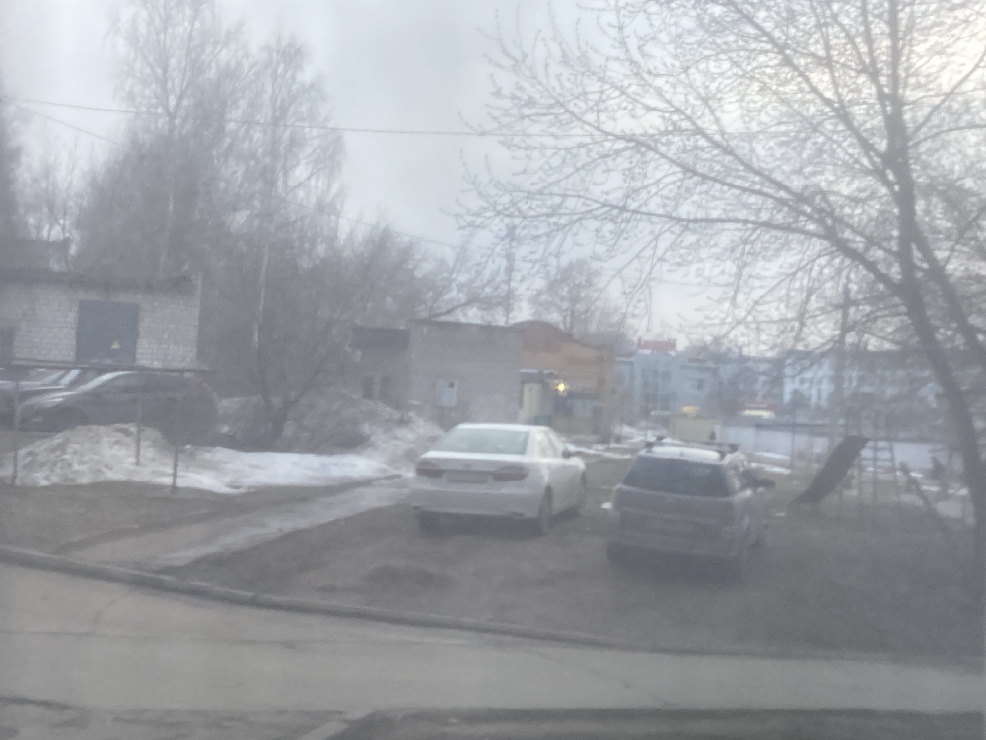 The image depicts a cloudy day in what appears to be a residential area in Chaikovsky, Russia. In the foreground, the lower portion of a window frame is visible, suggesting the photo was taken from inside a building. The immediate outdoor area consists of a dirt and gravel yard. Two cars are parked in the yard: a white sedan and a darker SUV. A pile of melting snow is present near a building to the left. Bare trees dominate the scene, adding to the overcast atmosphere. In the background, there are glimpses of other buildings, including some residential structures. A playground set can be seen to the right of the SUV.