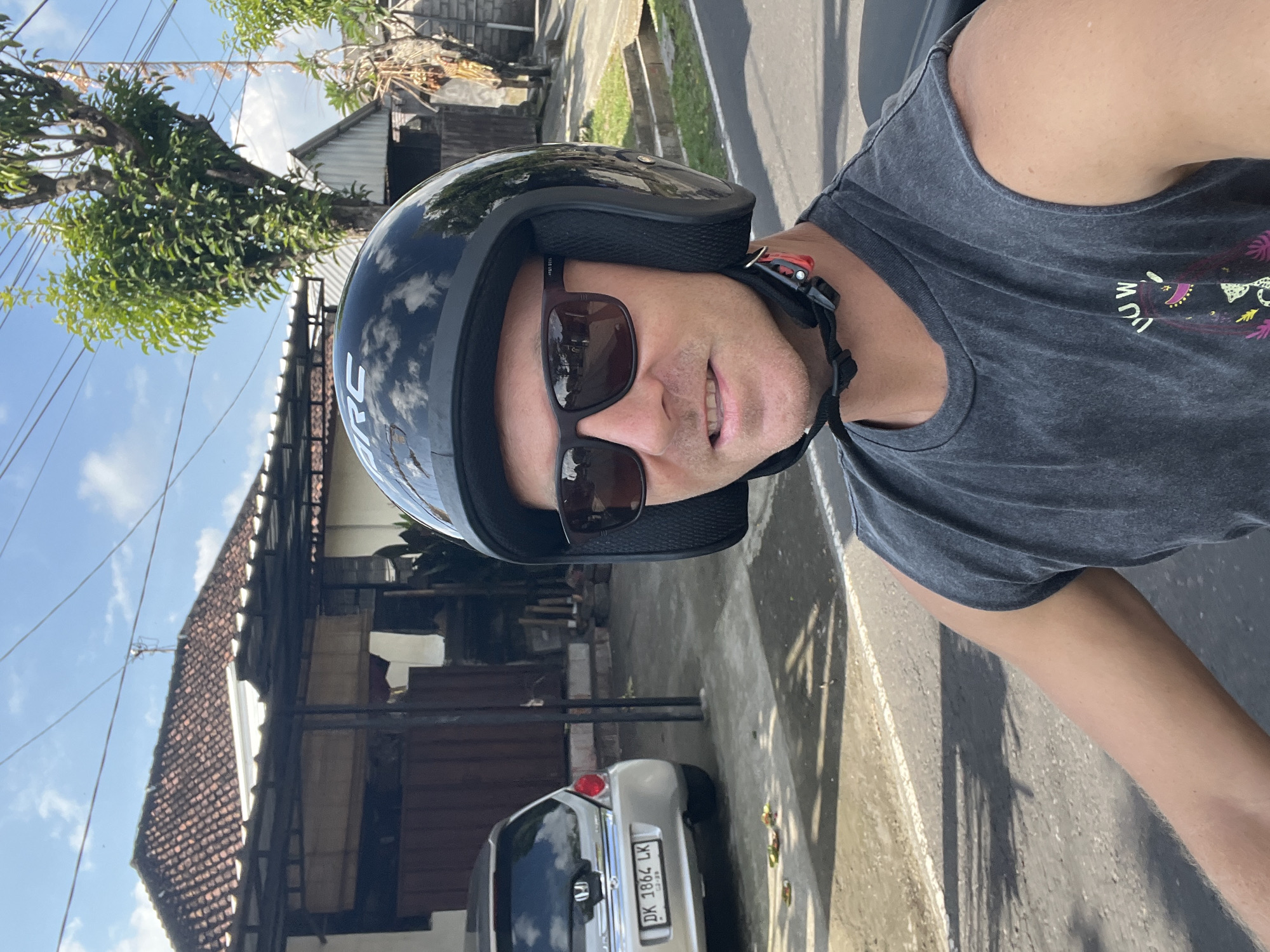 A man wearing a black motorcycle helmet and sunglasses is riding a motorcycle in Sukawati, Indonesia. He is wearing a dark grey tank top. A silver car with the license plate DK 1864 LK is parked on the side of the road. The background shows a house with a tiled roof, trees, and power lines. The day is sunny with a clear blue sky and some clouds.