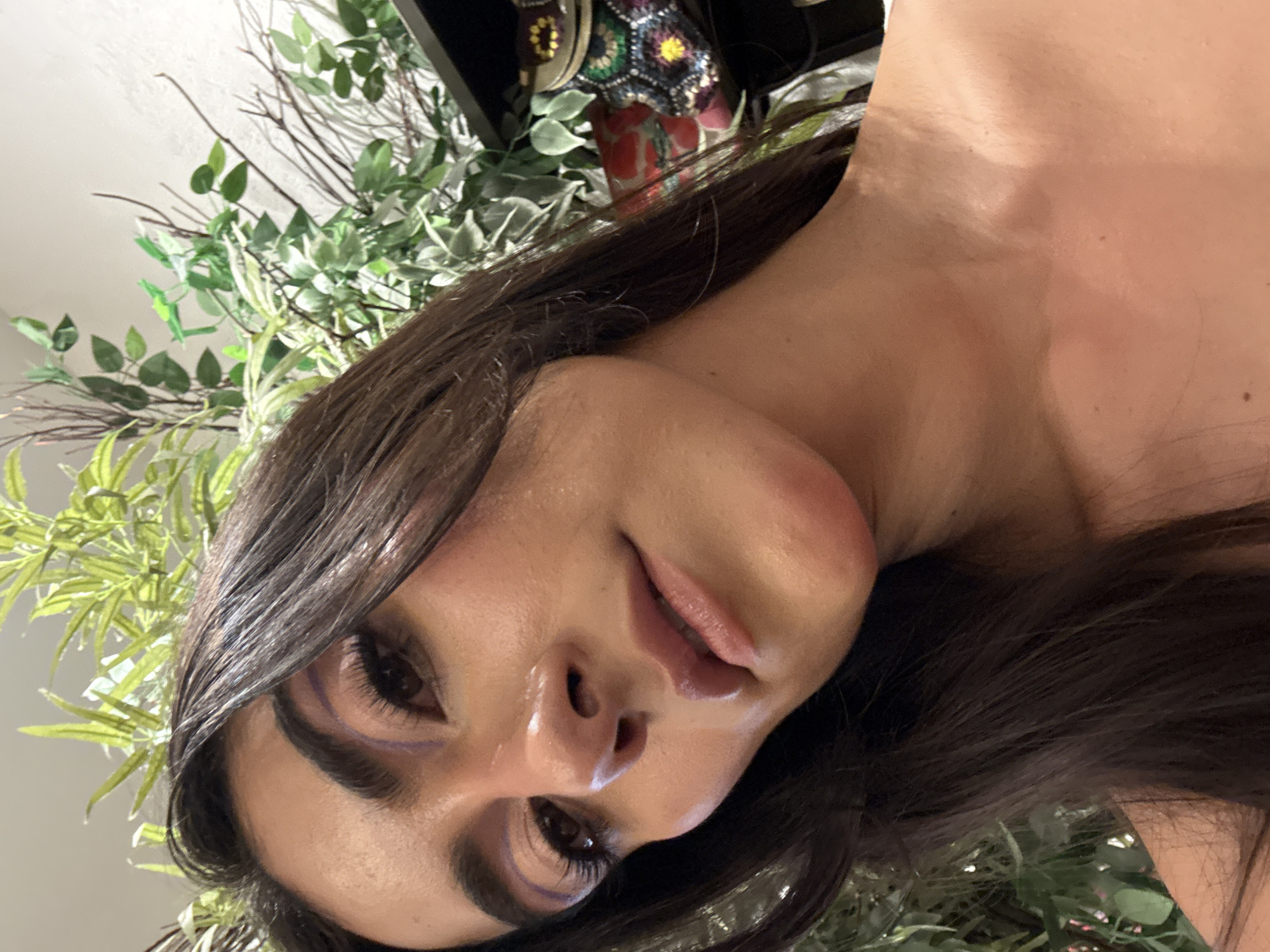 A close-up, rotated portrait of a woman with dark hair and fair skin is captured amidst foliage. The woman's face is angled towards the camera, showcasing her eyes with visible makeup. She is positioned against a backdrop of green plants and branches. In the upper part of the frame, behind her head, decorative objects and a portion of the ceiling are visible. There is no explicit information about the location being Domingo Arenas, Mexico, within the visible content of this image.