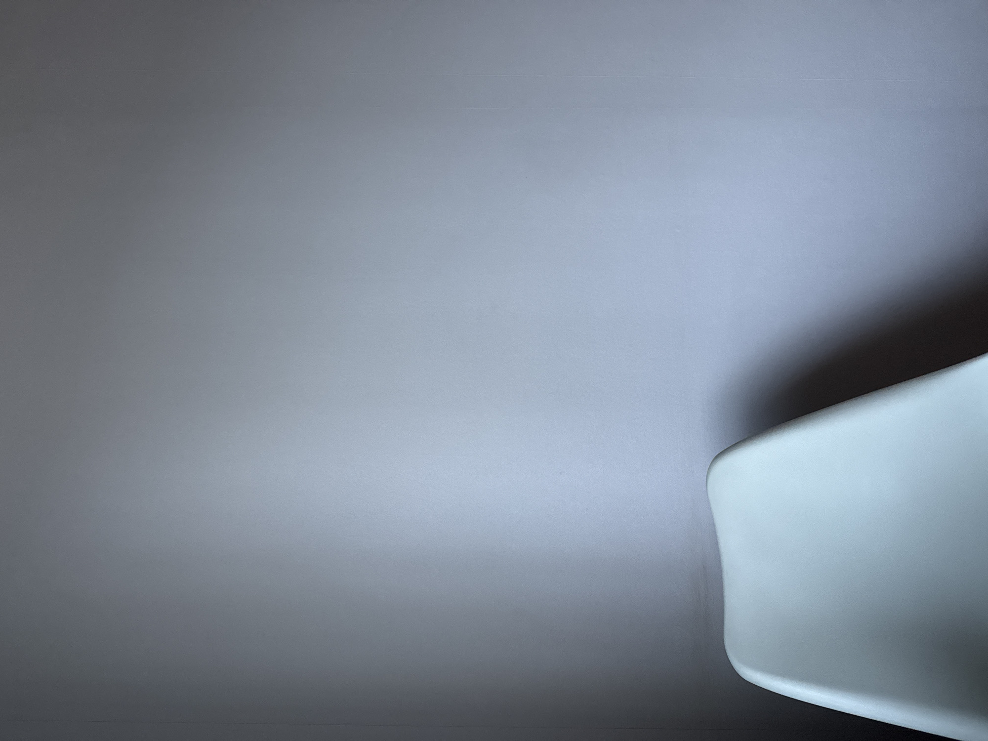 The image depicts a close-up view of a white object, possibly a chair, in the lower right corner, set against a background of a light gray wall. The scene appears to be indoors, and the lighting casts a soft gradient across the wall. The location is not specified.