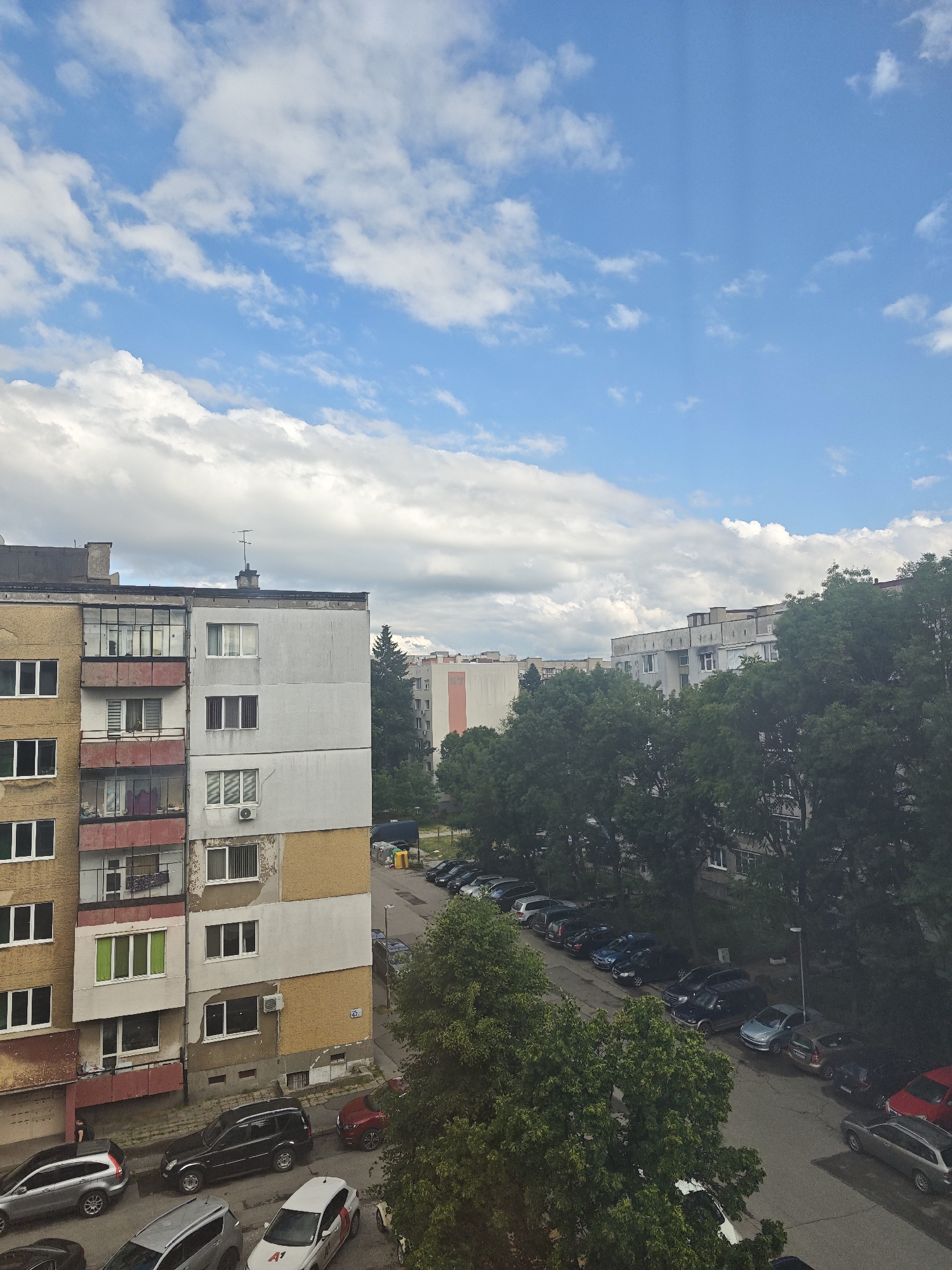 The image captures an elevated view of a typical residential urban area, likely from an apartment window or balcony. The scene depicts a densely built environment characterized by multi-story apartment blocks, lush green trees, and parked cars, consistent with a city like Sofia, Bulgaria.
In the foreground, a prominent apartment building occupies the left side, showcasing a mix of faded yellow and grey-white facades with visible signs of wear and tear, including peeling paint. It features numerous windows, some enclosed balconies with glass, and others with red railings. Air conditioning units are mounted below several windows. On the building's side, a small plaque displays the number "47" or "A7".
Abundant green deciduous trees fill the right side and midground, their dense foliage partially obscuring other buildings and a winding street below. This street is lined with numerous parked vehicles, including sedans, SUVs, and hatchbacks of various colors (silver, black, red, white, blue). In the immediate foreground, a silver SUV, a black SUV, a red car, and a white car are visible. The white car clearly shows a partial logo, "A1," which is a telecommunications company common in Bulgaria.
The sky above is bright and expansive, dominated by scattered white cumulus clouds against a clear blue background, suggesting fair weather during daytime, likely late morning or early afternoon. No strong shadows indicate the sun is high. There are no people actively engaged in noticeable activities within the frame, contributing to a tranquil, everyday urban atmosphere. The overall impression is one of ordinary life in a residential district during a pleasant day.