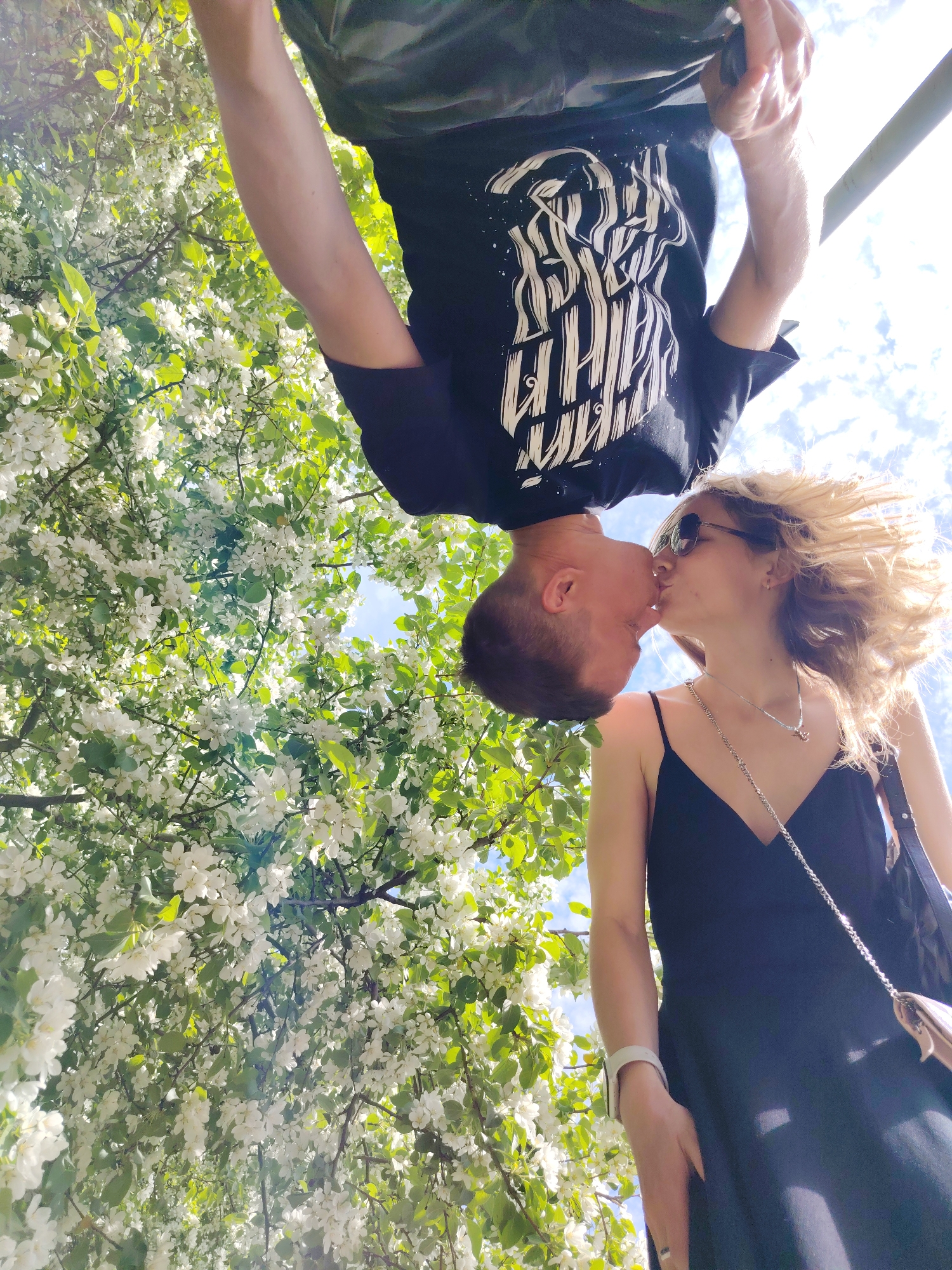 The photo depicts a man and a woman kissing beneath a flowering tree. The man is hanging upside down, his T-shirt featuring a stylized print. The woman is standing upright, wearing a black dress and sunglasses. The background features white blossoms of the tree and glimpses of the sky. The photo was taken in the city of Chaikovsky, Russia.