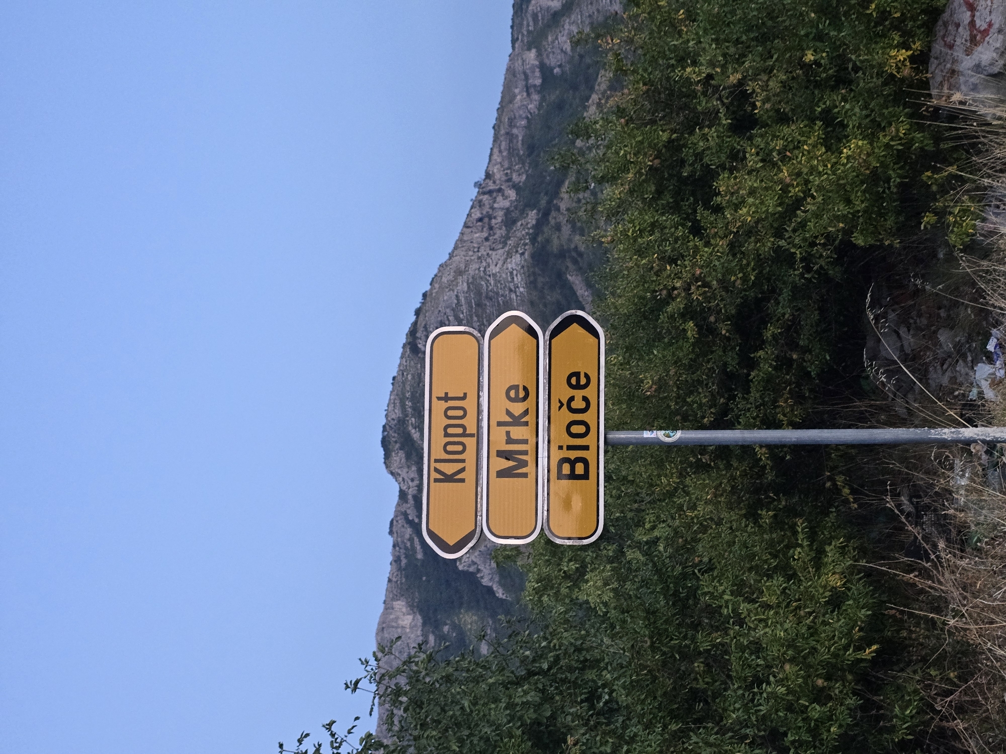 A directional signpost stands in front of a lush green bush and a rocky mountain under a clear blue sky. The signpost has three yellow signs with black lettering. The top sign reads "Klopot," the middle sign reads "Mrke," and the bottom sign reads "Bioče." The signs are pointing to the right. The mountain in the background is rugged and appears to be covered in sparse vegetation. The sky is a gradient of light blue, becoming darker towards the top. The foreground is filled with dry grass and some scattered debris. The overall scene suggests a rural or mountainous area, possibly a road leading to the locations indicated on the sign.
