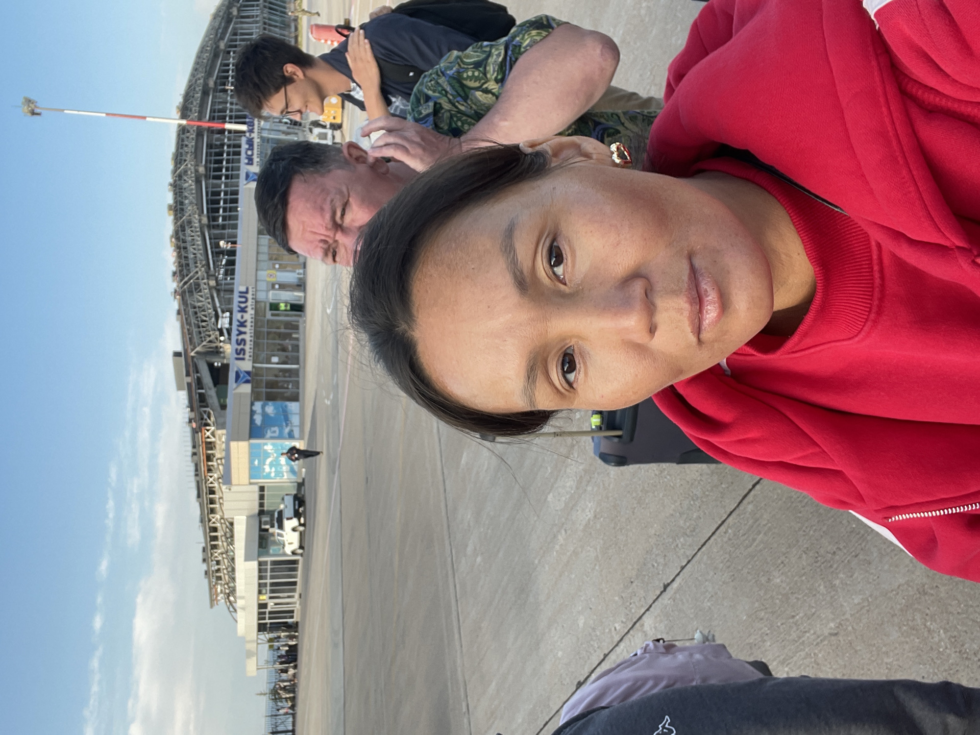 An outdoor, eye-level medium-shot image shows three individuals standing on an airport tarmac. The primary subject is a woman in the foreground, facing the camera. She has dark hair pulled back and is wearing a bright red hooded sweatshirt. She has a small, heart-shaped gold earring in her left ear. Behind her, a man with short, dark hair is visible, looking to his left with his face partially obscured. He is wearing a patterned green and blue shirt. To the right of the man, another individual, a man with glasses and dark hair, is looking to his right, with his left hand on the shoulder of the man in the green shirt. He is wearing a dark blue shirt and a backpack.
In the background, an airport terminal building is visible. The words "ISSYK-KUL" are prominently displayed on the building, with "International Airport" below it in English. The architecture of the building is modern, with a large, curved roof structure supported by exposed metal beams. A white pickup truck is parked near the entrance of the terminal. Other people are visible in the distance, as well as luggage. A red and white pole with a light fixture at the top stands near the terminal. The sky is clear with some clouds. The ground is paved with concrete. The location is identified as Issyk-Kul International Airport, near Choktal, Kyrgyzstan.