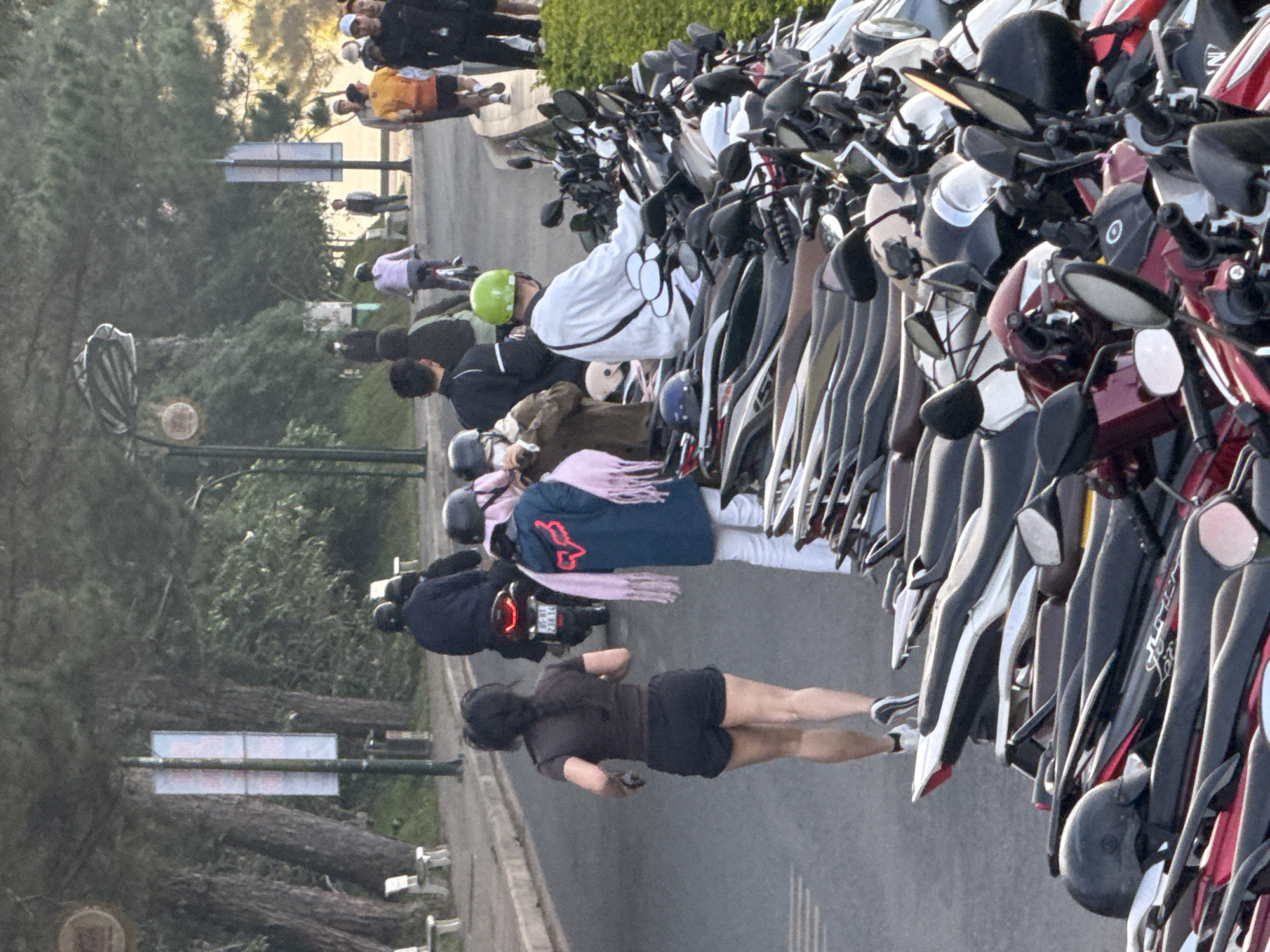 The image depicts a street scene in Da Lat, Vietnam, characterized by a row of parked motorcycles on the right side of the frame. On the left, a woman with dark hair in a braid is running away from the camera, wearing a dark t-shirt and black shorts. Behind her, several people are visible, some on motorcycles, and others walking. In the background, there are trees and a street with a sidewalk. The atmosphere suggests a busy urban environment, possibly during daylight hours given the lighting conditions.