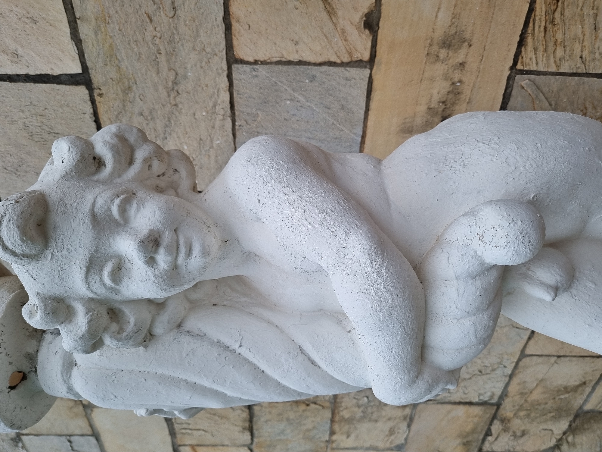 The image shows a close-up of a white statue, likely made of plaster or concrete, depicting a cherub or putto. The statue is positioned against a backdrop of a stone wall, which appears to be made of light brown and grey rectangular stones. The cherub is in a seated or embracing position, with its arms wrapped around a twisted pillar or column. Its face is visible, showing curly hair, closed eyes, and a slight smile. The statue shows some signs of wear, with peeling paint and visible textures. The lower portion of the statue reveals male genitalia. This statue is located in Laranjal Paulista, Brazil.