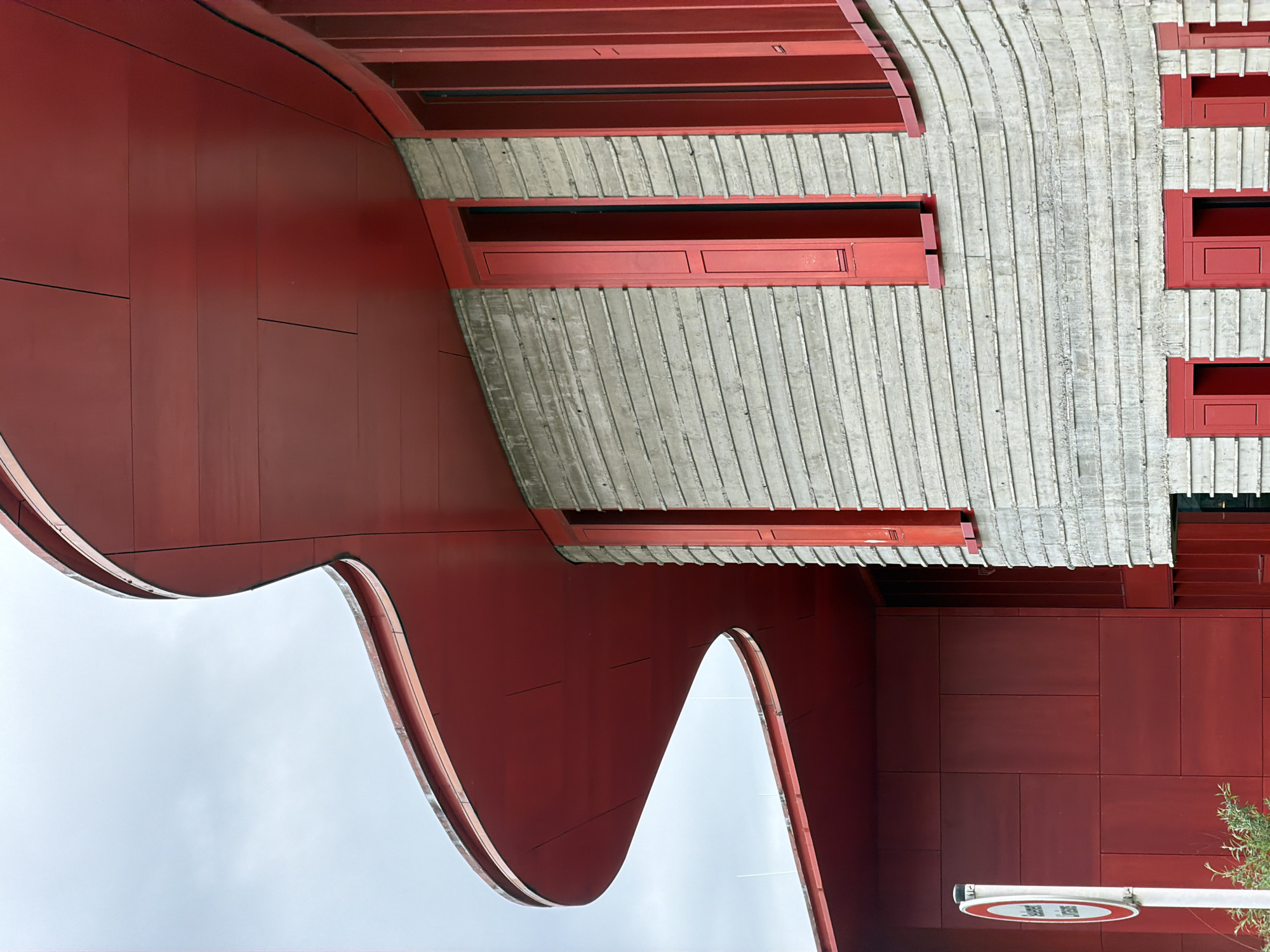 The image presents an architectural view of the Bridderhaus in Esch-sur-Alzette, Luxembourg. The structure showcases a combination of red panels and light-colored, ribbed concrete. A notable feature is the undulating red facade, contrasting with the linear texture of the concrete. Rectangular red window frames are visible within the concrete structure. In the lower right corner, a road sign and foliage are present. The sky, visible in the gaps of the facade, appears overcast.