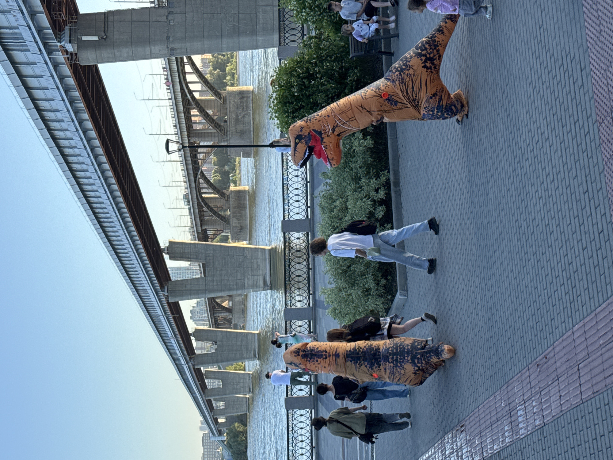 In Novosibirsk, Russia, a bright sunny day showcases the Ob River and a multi-span bridge. On the riverbank promenade, a person in a T-Rex costume walks towards the right, while another person in a similar costume stands to the left. Several other individuals, including a child, are walking or sitting on benches. The promenade is paved with brick and stone.