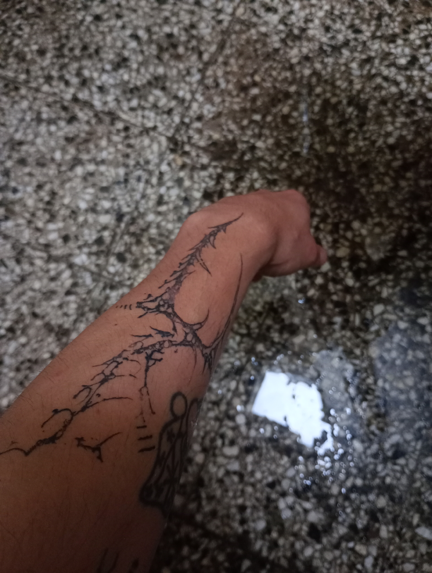 An adult human forearm is presented in a scene with a light, terrazzo-style floor covered partially in water. The forearm features a black ink tattoo design, which appears to be a combination of abstract geometric shapes and possibly stick-figure-like representations. The surrounding environment includes what seems to be a floor with a light-colored aggregate material, partially submerged in water, creating a wet or flooded appearance. The overall context provides no direct evidence to locate this image within the city of Buenos Aires, Argentina.