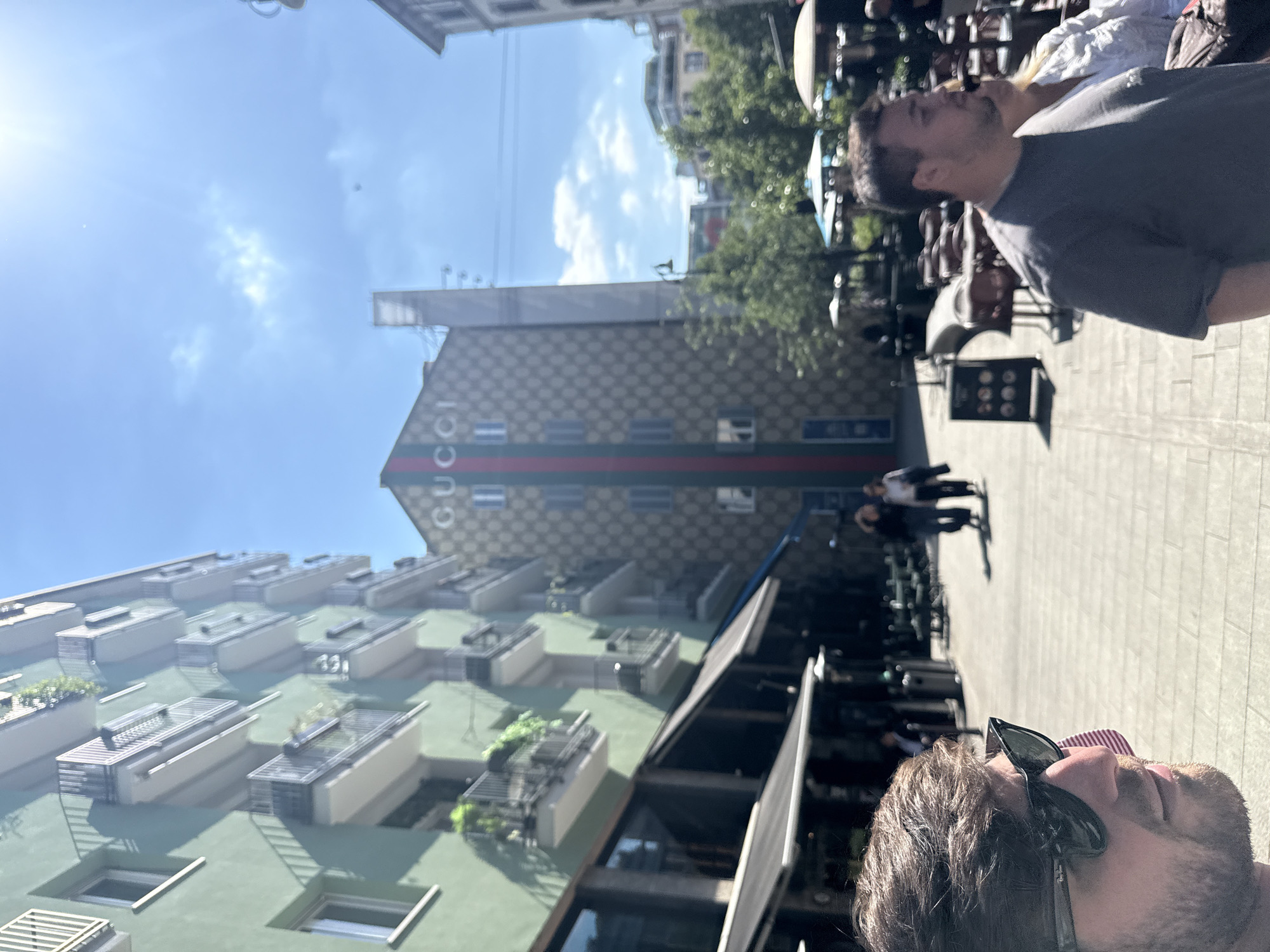 This is an outdoor street scene captured on a bright, sunny day in Milan, Italy. The image, oriented upright, features a paved street or sidewalk in the middle ground, populated by several individuals and outdoor furniture suggesting a cafe or restaurant setting.
In the foreground, to the left, is a close-up of a man's head and shoulders, looking upwards and slightly to the right. He has brown hair, a beard, and wears sunglasses atop his head. He is dressed in a striped shirt. Further back on the sidewalk, to the right, another man with dark hair and a beard is visible from the chest up, also looking skyward. Other blurry figures are scattered across the paved area, engaged in various activities like walking or standing.
The background is dominated by two prominent buildings under a clear blue sky with some scattered white clouds and a noticeable sun flare in the top right. To the left, a multi-story building with a green facade features numerous balconies, some adorned with plants or air conditioning units. Centrally located is a distinctive building with a patterned exterior, possibly tan and brown checkered, which clearly displays the word "GUCCI" vertically along its side, accented by a red and green stripe. Trees are also visible between some of the structures. The bright lighting suggests it is daytime, likely mid-morning or afternoon.