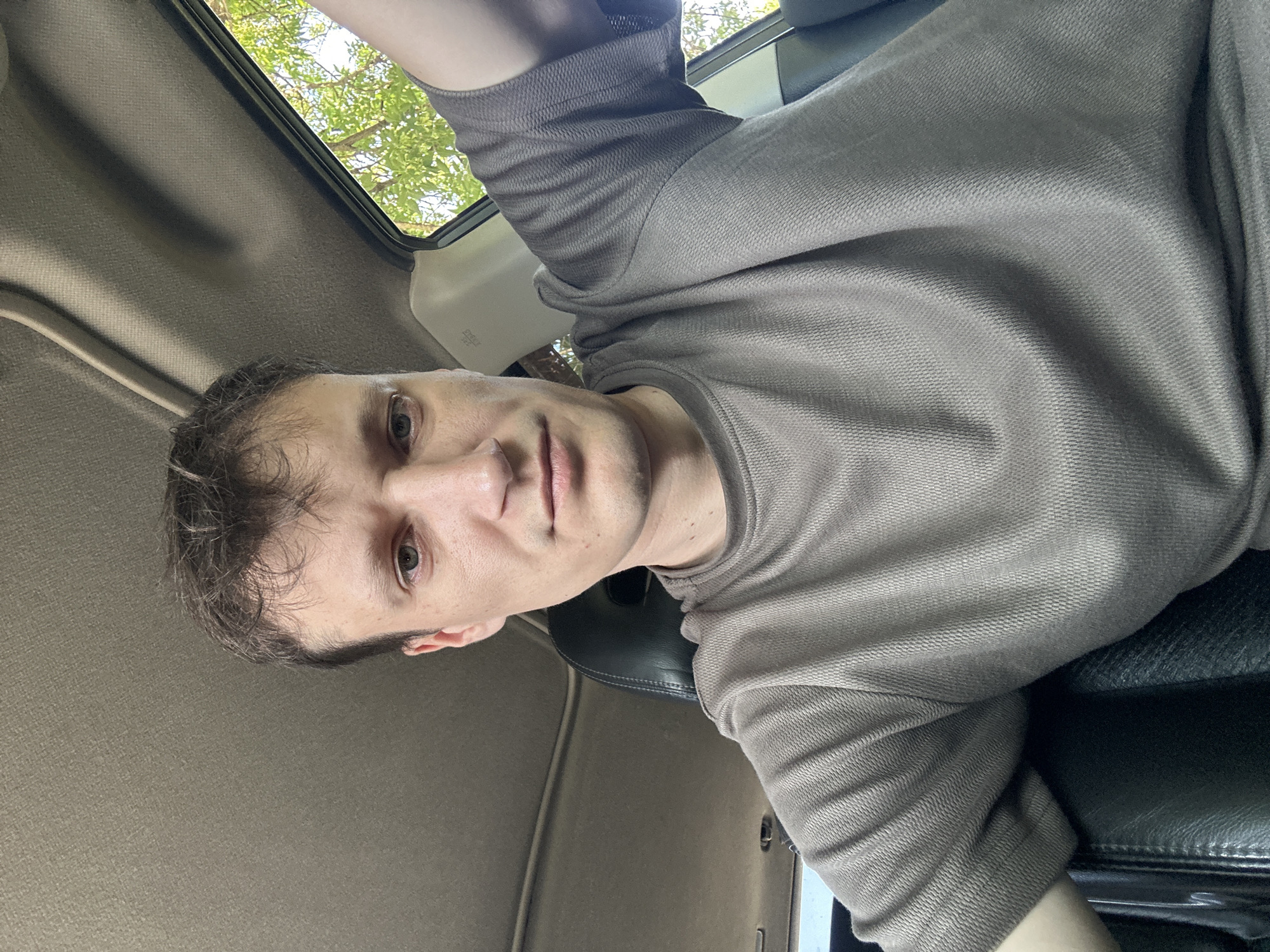 A medium shot captures a young man sitting in the driver's seat of a car, looking directly at the camera. He has short, dark brown hair and fair skin with some blemishes. He is wearing a gray, short-sleeved, textured t-shirt. The interior of the car is visible, with a dark gray headliner and a tan-colored sun visor. A black leather car seat is partially visible in the foreground. Through the open window on the right, lush green trees and foliage are visible under a bright sky, suggesting an outdoor location. The image appears to have been taken during the daytime, with natural light illuminating the scene. The location in Makiv, Ukraine, is not discernible from the visual information provided in the image.