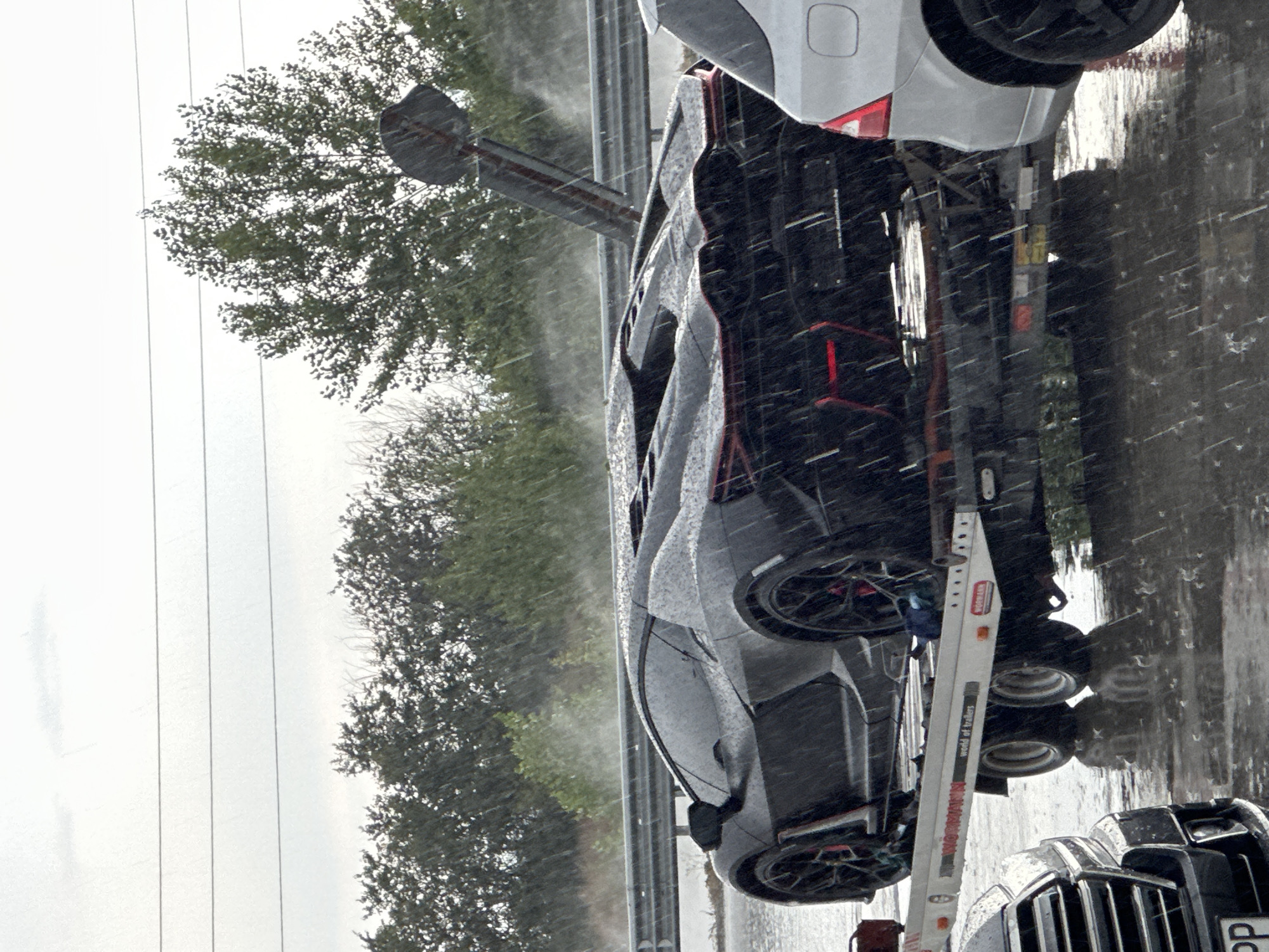 A black Lamborghini supercar is being transported on a trailer, wet from heavy rain. The trailer is on a flooded roadway, indicating a potential accident or unfortunate circumstance. Other vehicles are visible in the background, also affected by the weather. The overall scene depicts a challenging and possibly hazardous driving environment, with the luxurious sports car exposed to the elements. The location is identified as Polsko Padarevo, Bulgaria.