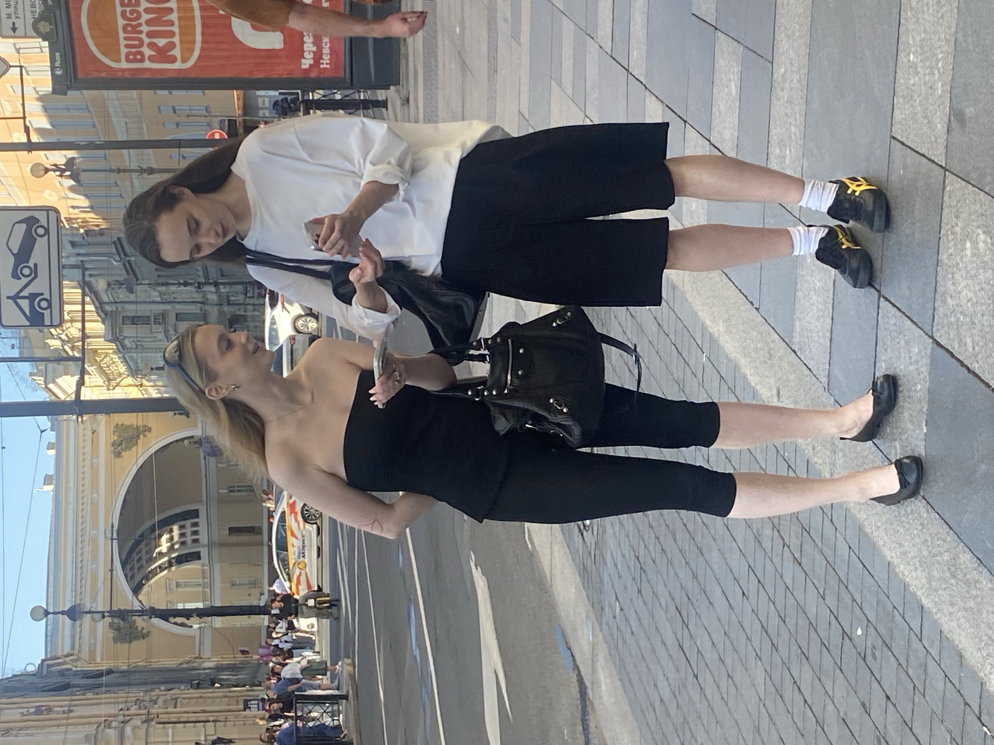 Two young women are standing on a paved sidewalk in Saint Petersburg, Russia, near the Palace Square. The woman on the left, wearing a black strapless top and black capri leggings, is holding a smartphone in her right hand and looking down at it. She has blonde hair with a few strands falling forward. A large black handbag is slung over her left shoulder. The woman on the right, with long brown hair, is wearing a white long-sleeved shirt and black culottes. She is also holding a smartphone in her hands and looking at it. She has white socks and black and yellow sneakers. In the background, a yellow car with Russian license plates is driving on the street, and a large arched building, possibly the Main Staff Building, is visible. Several other pedestrians are in the background, and a Burger King advertisement is on a red billboard to the right. The sky is clear and blue, indicating a sunny day.