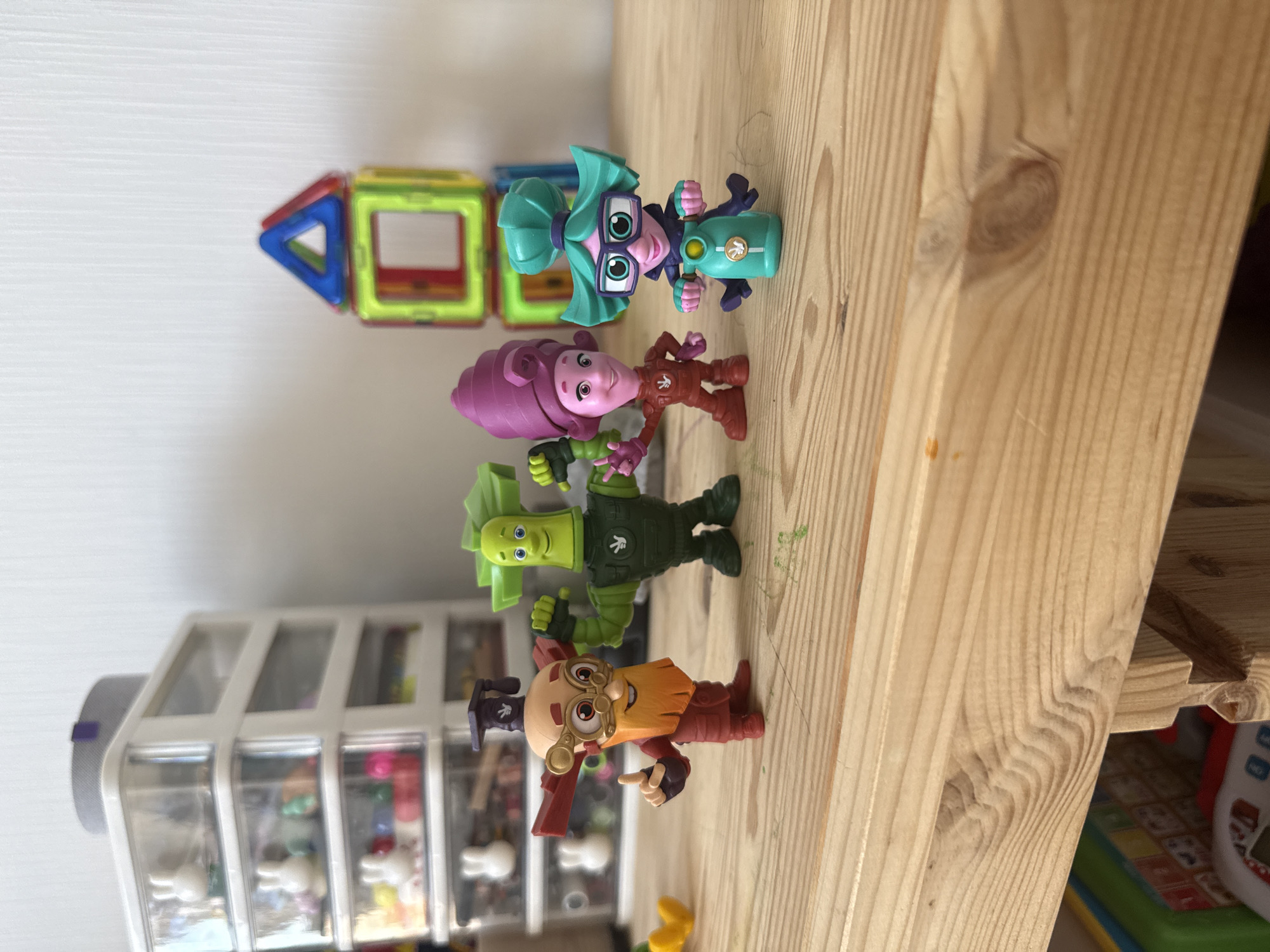 Four animated figurines stand on a wooden surface. From left to right, the first figurine has a ginger beard, goggles, and a graduation cap on its head. It is wearing a red outfit and has red wings. The second figurine is green with large, spiky hair. It is wearing a dark green suit. The third figurine has purple hair and is wearing a red, form-fitting outfit. The fourth figurine has green hair and is riding a turquoise scooter. It is wearing a dark outfit and large glasses. Behind the figurines, a white storage unit with clear drawers is visible, containing small white rabbit figures and colorful objects. To the right of the storage unit, a colorful magnetic building toy is assembled into a house shape. The wooden surface appears to be a shelf or table. The background is a plain white wall. The image is a still photograph taken in a room.
