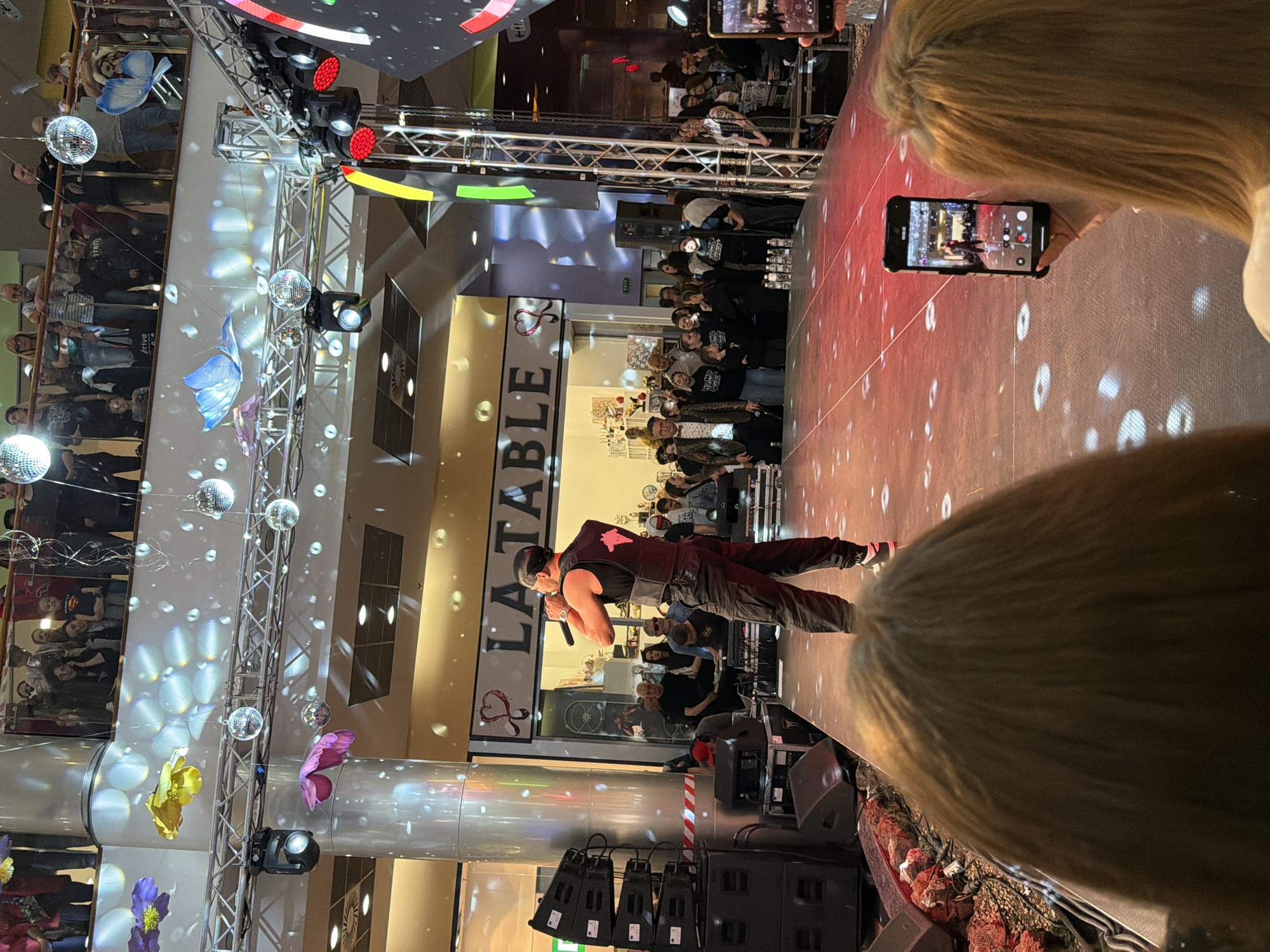 This image captures a lively indoor performance event in what appears to be a multi-level shopping mall or large public venue in Pleven, Bulgaria.
At the center of the scene is a male performer on stage, dressed in dark attire including a sleeveless top with a pink logo (resembling the Kappa brand) and a backward cap. He is holding a microphone and appears to be singing or addressing the audience. The stage itself is red and illuminated with numerous white circular and star-like light projections, suggesting a disco or party atmosphere. Stage lighting fixtures, including red spotlights, are visible on metal truss structures, along with large speakers on the stage floor.
An engaged audience surrounds the stage, both on the ground level directly in front of the performer and from a higher balcony or walkway on the left side of the frame. Many audience members are holding up their mobile phones to record the event. In the immediate foreground, the back of a blonde-haired person's head is visible, partially obscuring the bottom right of the image, and another person is seen holding a smartphone, filming the stage.
The venue is decorated with multiple reflective disco balls hanging from overhead structures and large, colorful artificial butterflies in shades of blue, yellow, and pink. In the background, a storefront sign partially reads "LA TABLE," accompanied by a heart-shaped logo. The setting, with its artificial lighting and festive decorations, indicates an evening or nighttime event.