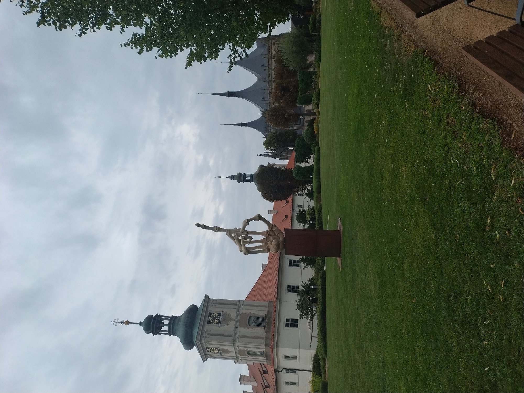 In Kutná Hora, Czechia, a sculpture resembling a skeletal hand giving the middle finger stands prominently on a metal pedestal in a manicured lawn. The sculpture is positioned in the foreground, to the left of center. Behind it, a large white building with a red tiled roof features a prominent clock tower topped with a green spire. To the right of this building, and further in the background, is a striking Gothic church with multiple spires and a dark, patterned roof. The sky is overcast with a grey, cloudy texture. Trees with green foliage are visible on the right side of the image, and a path with wooden benches runs along the far right edge of the frame.