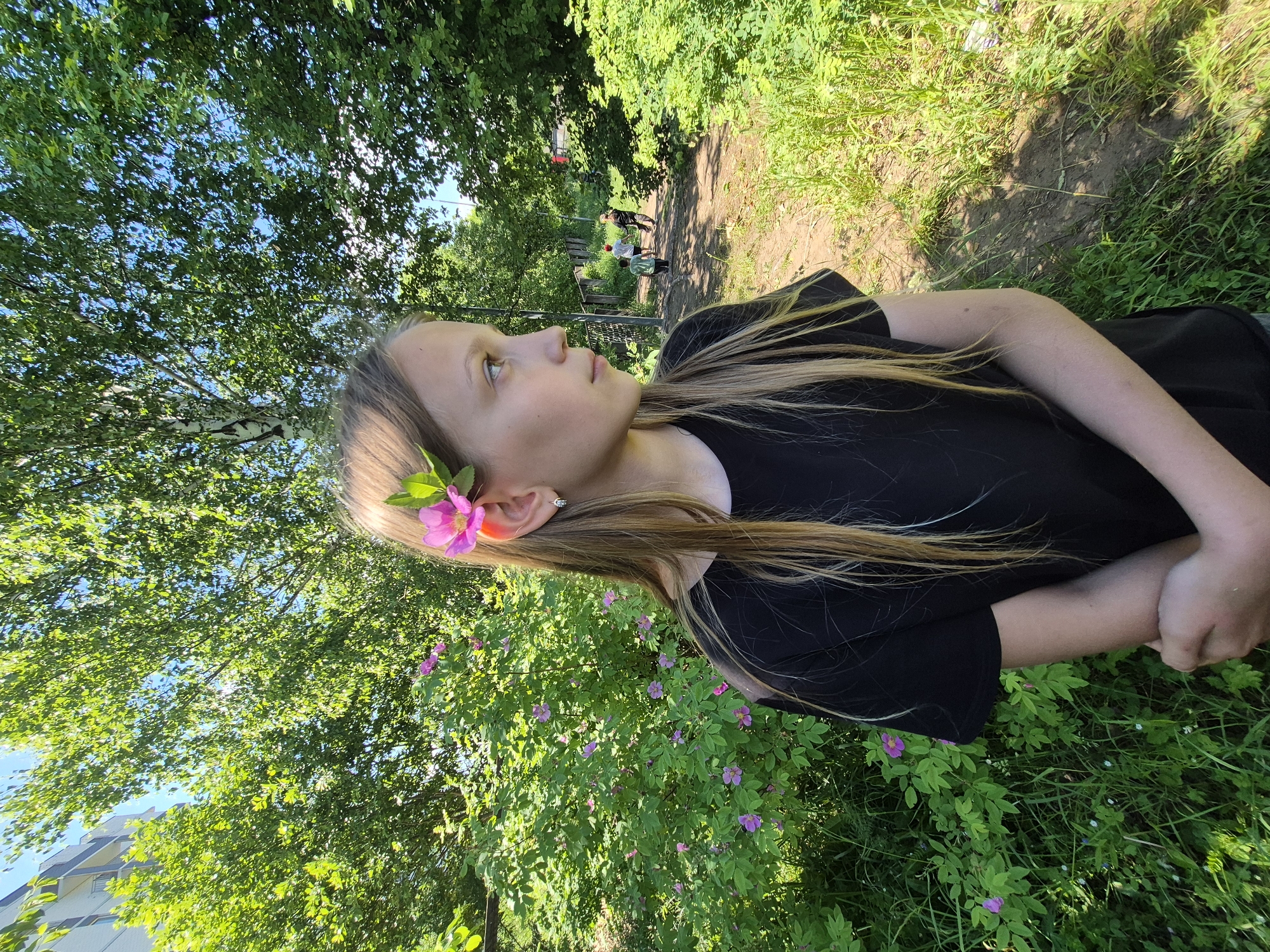 A young girl with long blonde hair is standing in an outdoor setting, with a pink flower tucked behind her right ear. She is wearing a black t-shirt. Behind her, a lush green bush with pink flowers is visible, along with trees and a path where two children and an adult are walking in the distance. The area appears to be a park or a residential area with a building visible to the left. The scene suggests a sunny day in Chaikovsky, Russia.