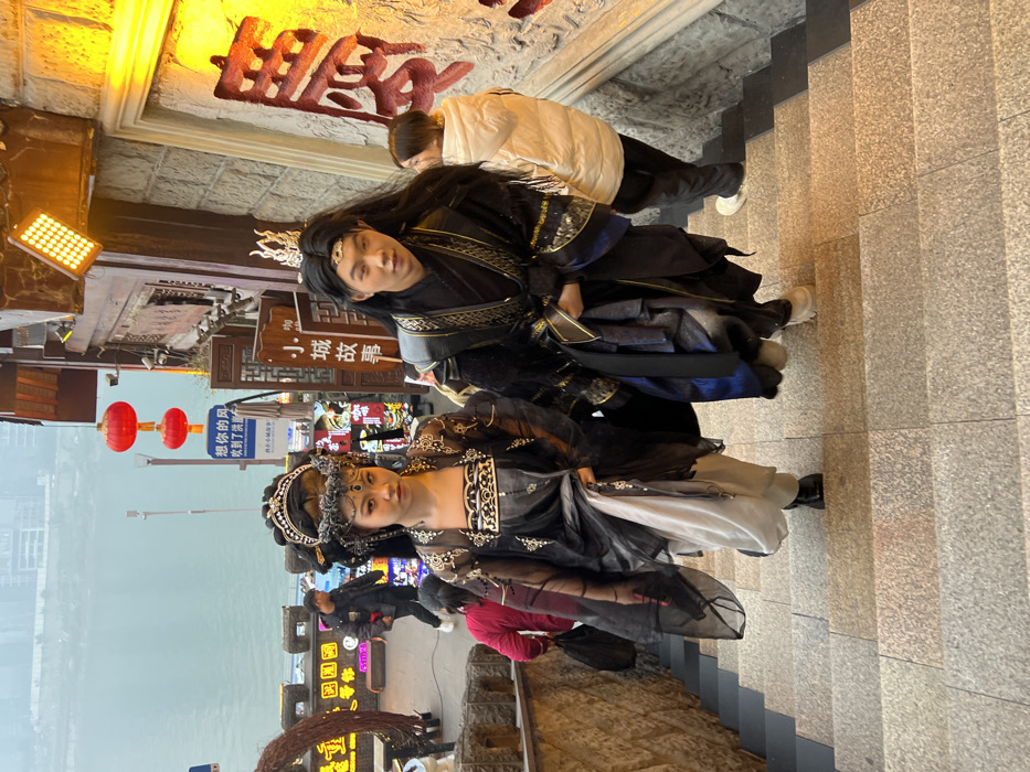 Two individuals in elaborate traditional Chinese attire descend a stone staircase in Yuzhong District, Chongqing, China, likely within the Hongya Cave complex. The female subject on the left wears a multi-layered black and grey ensemble with a white underskirt, featuring intricate embroidery and a large, ornate dark headdress with silver accents. She has distinct eye makeup and looks directly forward. To her right, a male subject, with long dark hair, wears a black and blue robe with gold trim and a white, pointed crown. He holds the fabric of his robe and looks slightly to his right.
They are positioned centrally on a wide, light-grey stone staircase. To their immediate right, a stone wall with a prominent illuminated sign bearing red Chinese characters is visible. Above the sign, a bright yellow LED floodlight is mounted. To their left, a traditional-style building facade with decorative wooden elements and hanging red Chinese lanterns is present. A blue sign with white Chinese characters is visible in the mid-distance on the left. In the background, beyond a stone railing, a wide body of water, presumably the Jialing River or Yangtze River, stretches towards distant urban buildings under an overcast sky. Other people are visible in the scene: a person in a white puffer jacket is descending the stairs behind the main subjects, and another individual in a dark jacket walks further back near the waterfront. The overall environment suggests a bustling cultural or tourist area with historic architectural influences.