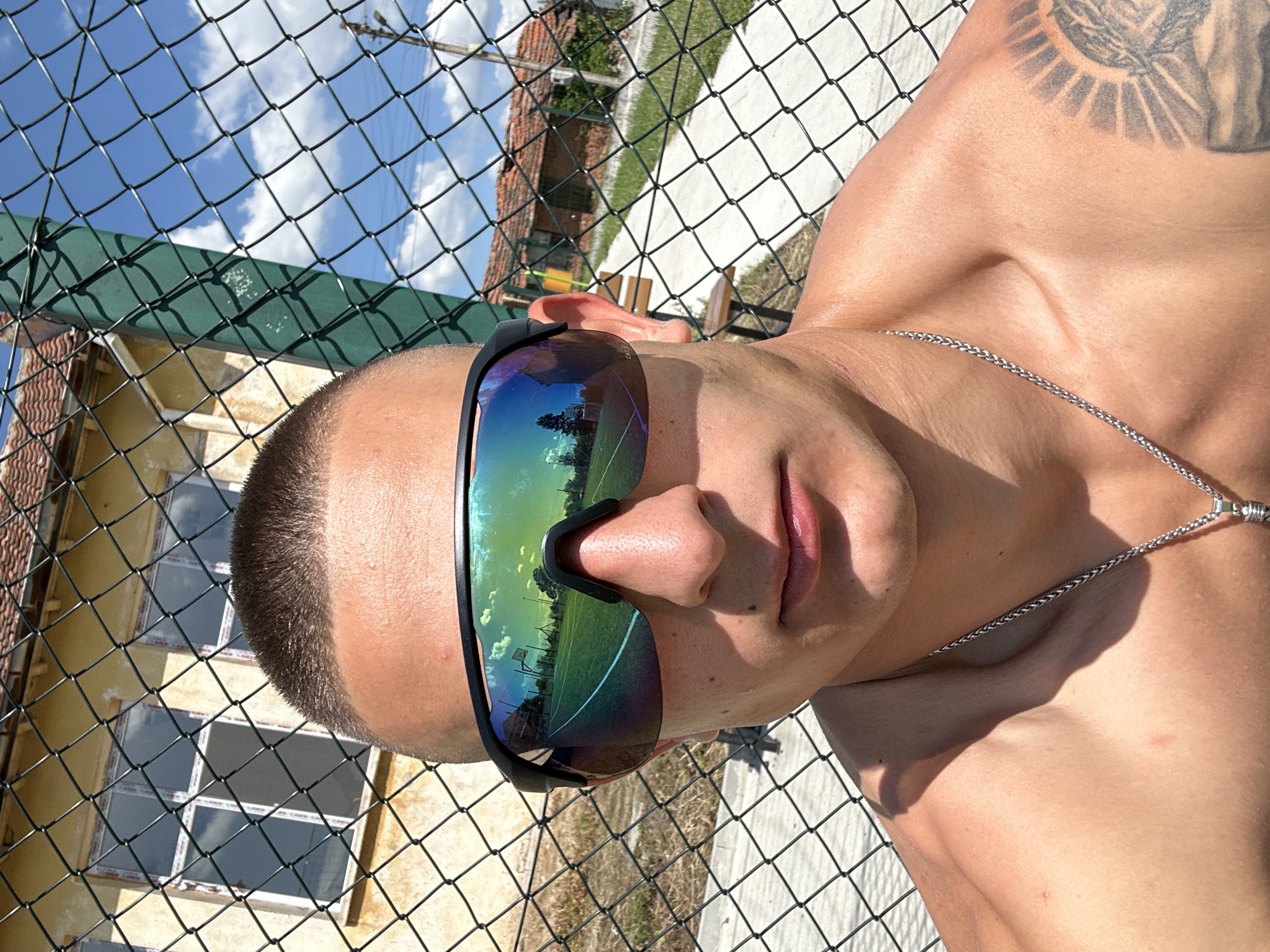 The image shows a man lying down, shirtless, facing upwards, with a black chain-link fence separating him from a building and a view of the sky. The man is wearing sunglasses with a reflective surface showing a green field and sky. The man's head is close to the fence. There is a tattoo on his upper right shoulder. Behind the fence, a building is partially visible with white-framed windows. The sky is blue with some clouds. The background suggests an outdoor area. The location is indicated to be Kalchevo, Bulgaria.