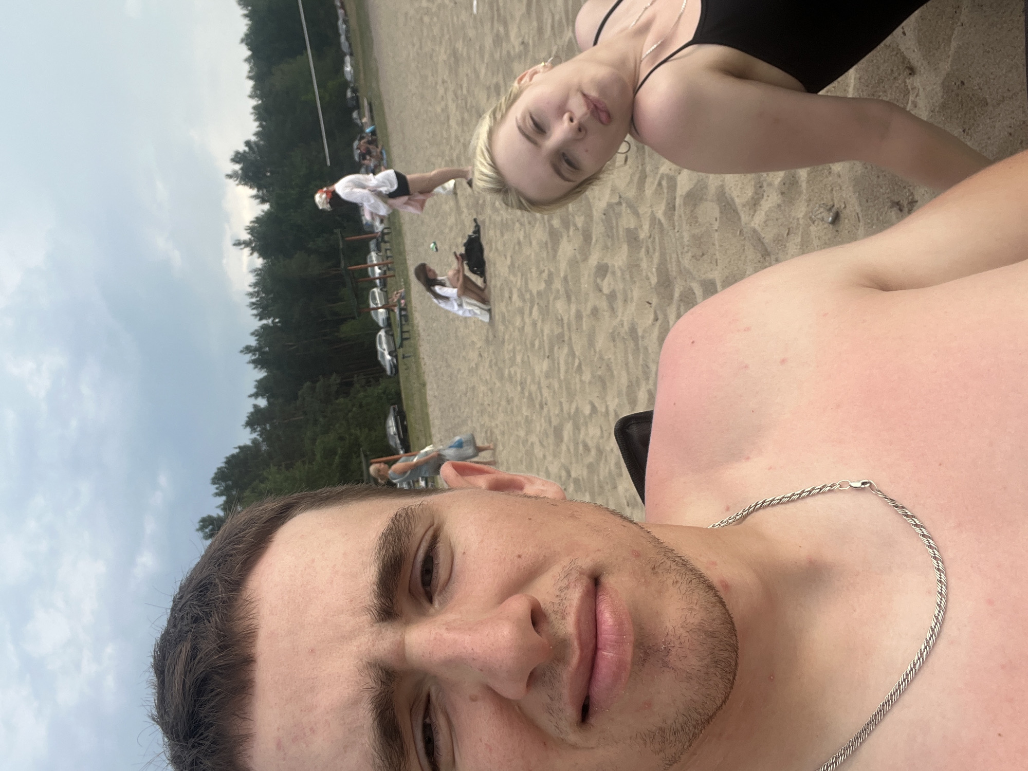 A selfie is taken on a sandy beach with trees and a cloudy sky in the background. The photo is close-up, with the face of a young man in the foreground, looking directly at the camera. He has short brown hair and is wearing a silver chain. To his right, and slightly further back, is a young woman lying down and looking away from the camera with her tongue sticking out. She is wearing a black swimsuit. In the background, other people are visible walking and sitting on the beach, and cars are parked near the treeline. There appears to be a volleyball net set up in the distance. The location is stated to be Загародскі сельскі Савет, Belarus.