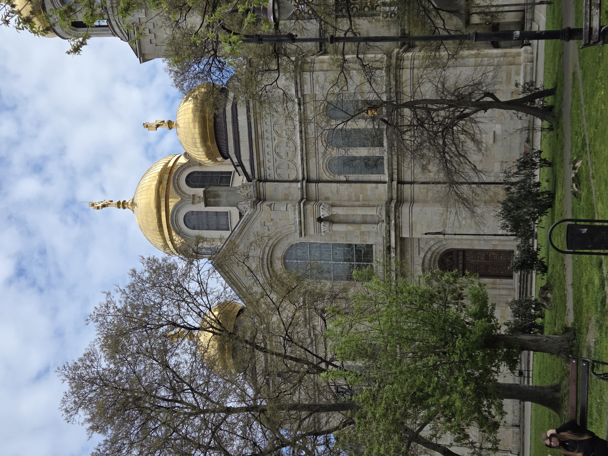 This image captures the exterior of the Dormition of the Theotokos Cathedral in Varna, Bulgaria, presented from a slightly low-angle perspective, emphasizing its scale and architectural details. The cathedral features a grand, light-colored stone facade with multiple arched windows, some stained or frosted, and intricate decorative carvings. Several prominent golden domes, each topped with a cross, rise towards the sky, with two being particularly dominant in the composition. Another smaller, unadorned dome is visible on a side tower.
The building is set within a green, park-like environment. Lush green grass covers the foreground and extends to the sides, with a paved path visible on the right. Several trees are scattered around the cathedral; some are still largely bare, while others display fresh, vibrant green leaves, suggesting early spring. A tall, black street lamp stands near the left side of the cathedral, and a dark park bench and a partial view of a trash can are visible on the right.
One person is partially visible on the far right, seated or standing behind the park bench. They have long, light-colored hair, wear glasses, and are dressed in dark clothing.
The scene is illuminated by natural daylight under a sky that is a mix of bright blue with scattered white clouds. The overall impression is that of a calm day at a significant religious and historical landmark. No legible text is visible in the image.