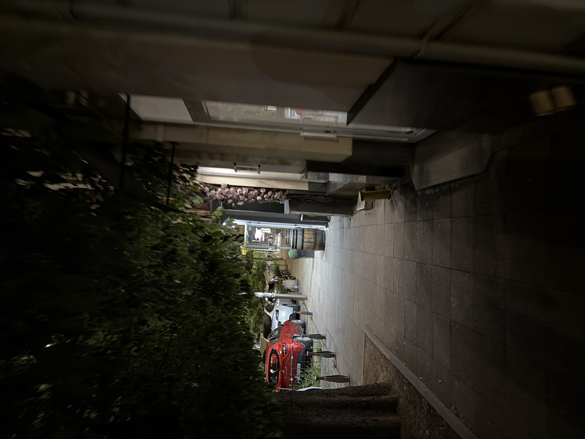The image depicts a sidewalk scene at night in Stara Zagora, Bulgaria. A red car is parked on the right side of the street, along with a white car and several others further down. A large tree with dark green foliage overhangs the sidewalk from the left. The sidewalk is paved with square tiles and is bordered by a curb. To the right of the sidewalk, there is a building with a storefront featuring a glass door and windows. A wooden barrel and a green planter are placed on the sidewalk near the building. The overall lighting suggests it is nighttime, with some artificial lights illuminating parts of the scene.