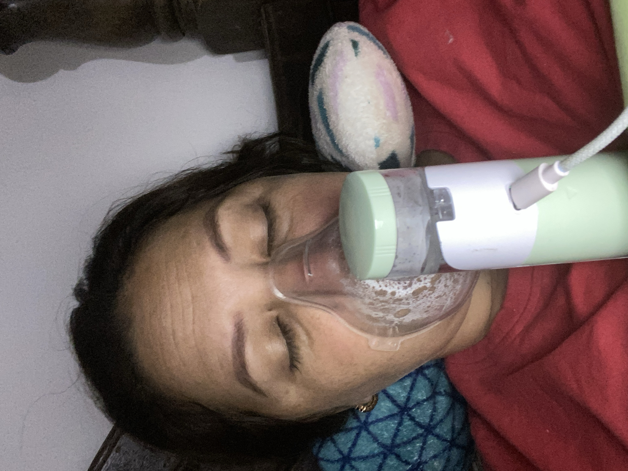 A person's face is partially obscured by a clear mask connected to a nebulizer. The person's eyes are closed, and their head is tilted back. The nebulizer is a green and white device with a clear chamber filled with liquid. A cord is plugged into the top of the nebulizer. The person is lying down, and a red garment and a blue and white patterned blanket are visible. A fuzzy object with pastel colors is behind the person's head. The scene appears to be indoors.