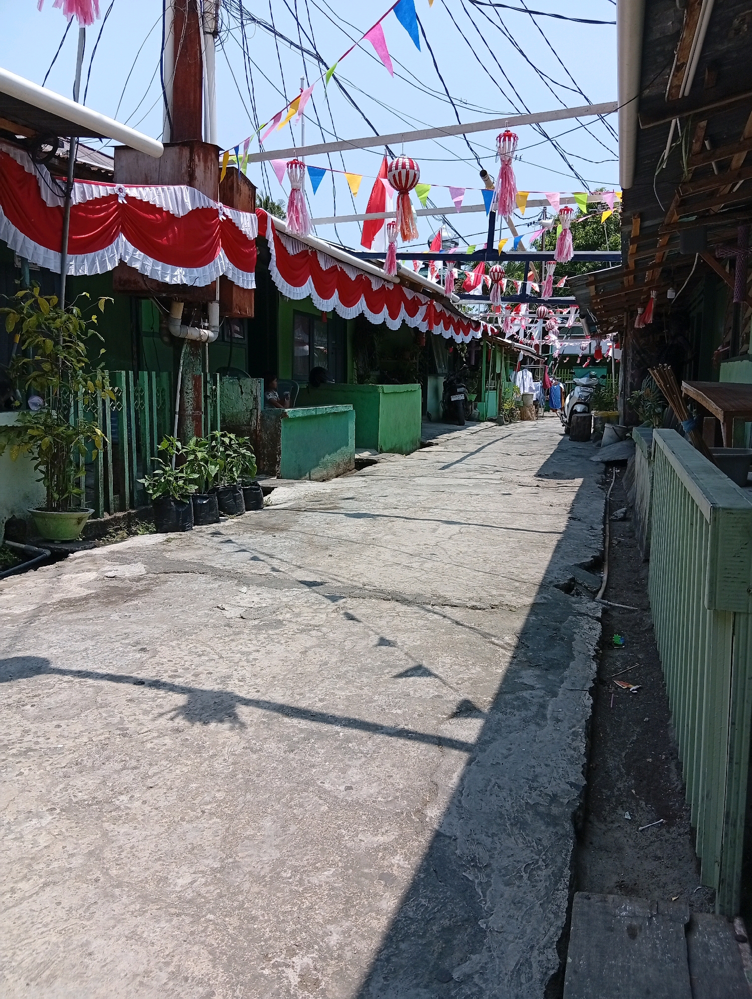 The image depicts a narrow, paved alleyway in a residential area, possibly in the city of Manado, Indonesia, as suggested by the festive decorations. The alley is lined with single-story buildings on both sides, painted in a muted green color. The buildings appear to be closely situated, creating an intimate urban environment.
The alleyway itself is constructed from concrete, showing signs of wear and tear, with some cracks and stains visible. The overhead view reveals a dense network of electrical wires crisscrossing the sky, indicative of an urban setting. The scene is further enlivened by numerous decorations, including red and white fabric draped along the eaves of the buildings, and colorful triangular flags and lanterns strung across the alley. These decorations suggest a festive occasion, such as a national holiday or a local celebration.
On the left side of the alley, several potted plants and small trees are visible, adding a touch of greenery to the otherwise utilitarian landscape. A tall, rusty metal pipe, possibly part of an exhaust or ventilation system, stands prominently near the left side of the alley.
On the right side of the alley, a light green metal fence runs along the edge of a property, bordering a drainage ditch filled with dark soil and some debris. Various objects, including what appear to be wooden planks, are also present near the fence.
In the distance, a few people are seen walking along the alley, and a motorcycle is parked on the left side. The overall impression is that of a lively, community-oriented street, decorated for a special event, set within a dense urban environment in Manado, Indonesia.