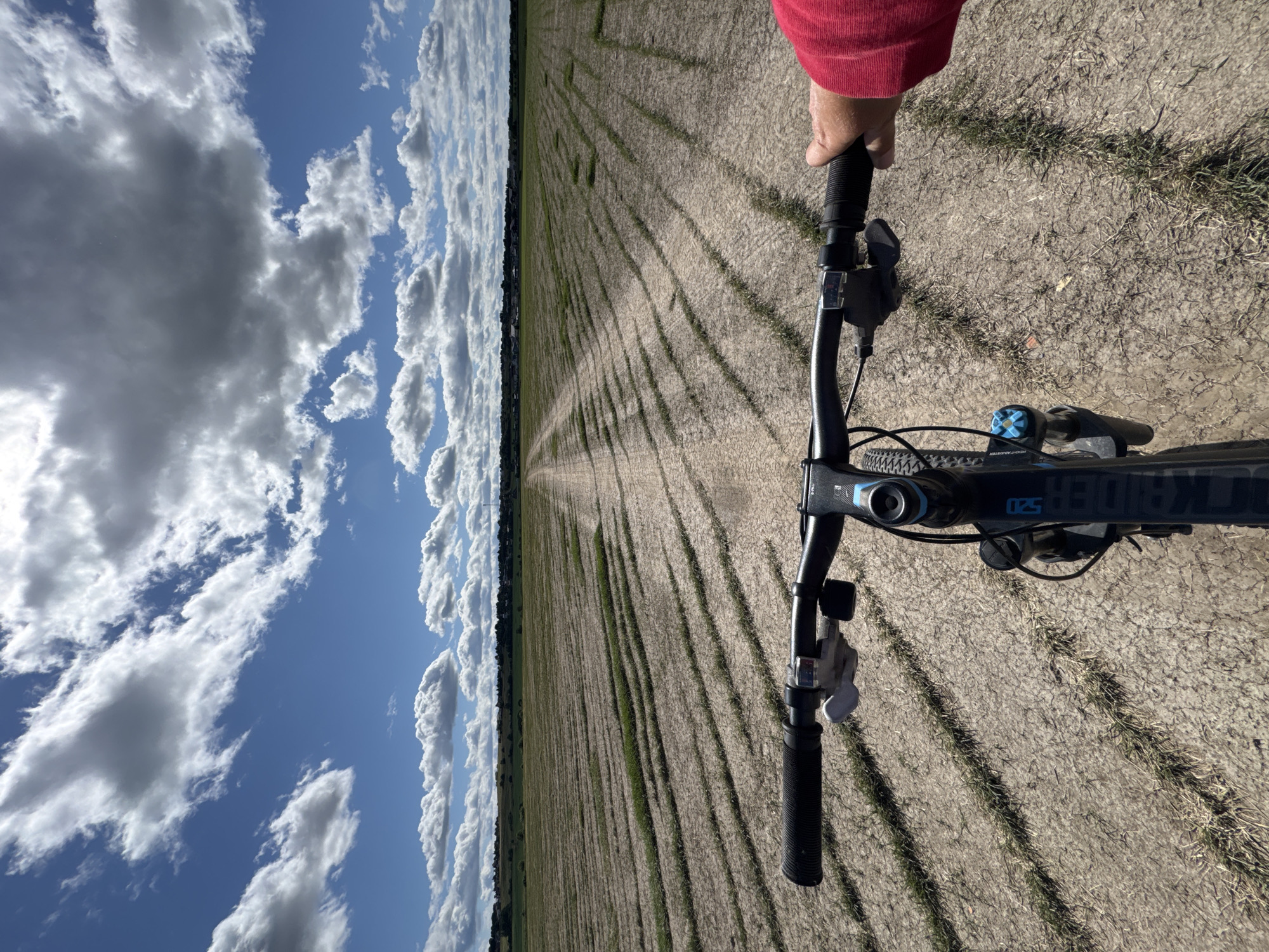A first-person perspective shot from a bicycle shows a dirt path leading through a dry, harvested agricultural field with rows of stubble. The field extends to the horizon where a distant tree line and scattered buildings are visible. The sky is a bright blue with scattered, fluffy white clouds. The handlebars and front wheel of the bicycle are in the foreground, with a person's right hand wearing a red sleeve gripping the right handlebar. The scene is in Kempelevo, Russia, under a partly cloudy sky.