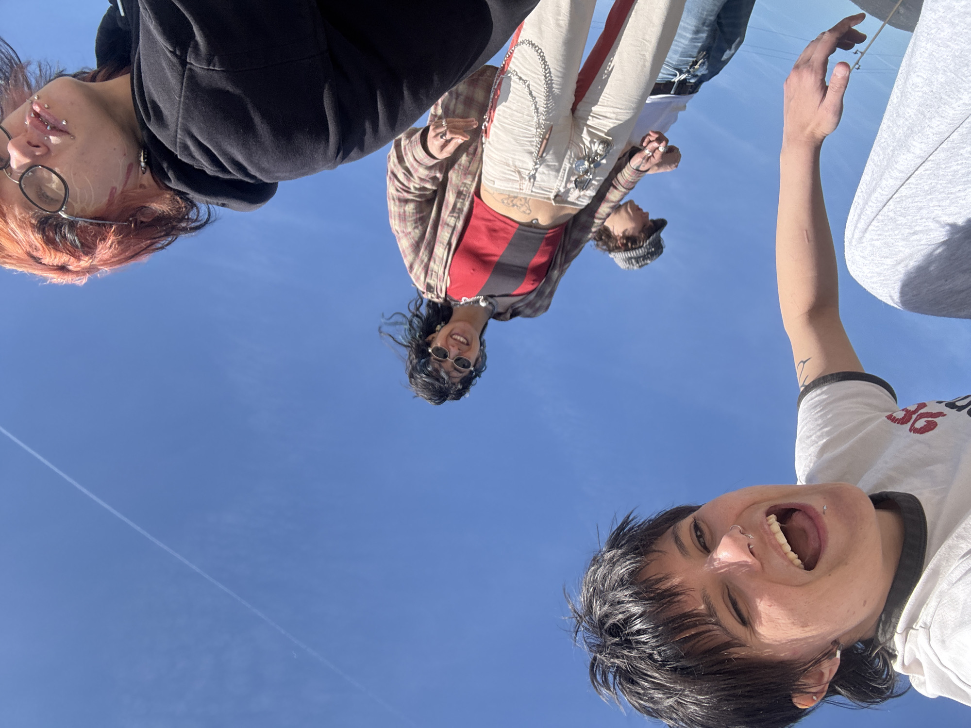 A low-angle shot depicts five individuals in what appears to be an outdoor setting, with a clear blue sky as the backdrop, and a contrail from a jet. The perspective is from below, looking up at the subjects.
One individual to the left of the frame is wearing glasses, with visible piercings on the face and red-tinted hair. Another is wearing a dark hoodie.
In the center, a person wears sunglasses, a plaid shirt, and light-colored pants with a chain and a decorative patch. Behind them is an individual wearing a beanie hat.
To the right of the frame, an arm is outstretched and one person is close to the camera with an open mouth.
The location is unknown.