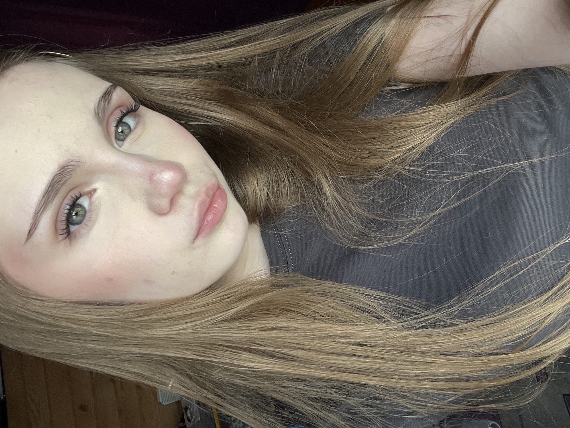 The image is a close-up, head and shoulders portrait of a young Caucasian female with long, straight, light brown hair. She has green eyes and is wearing a dark grey t-shirt. Her skin appears fair with some light blush on her cheeks. She is looking slightly off-camera to her left. The background consists of a wooden wall on the left and a dark purple curtain on the right. There is no indication of Ashkadar, Russia, or any specific urban environment within the image.