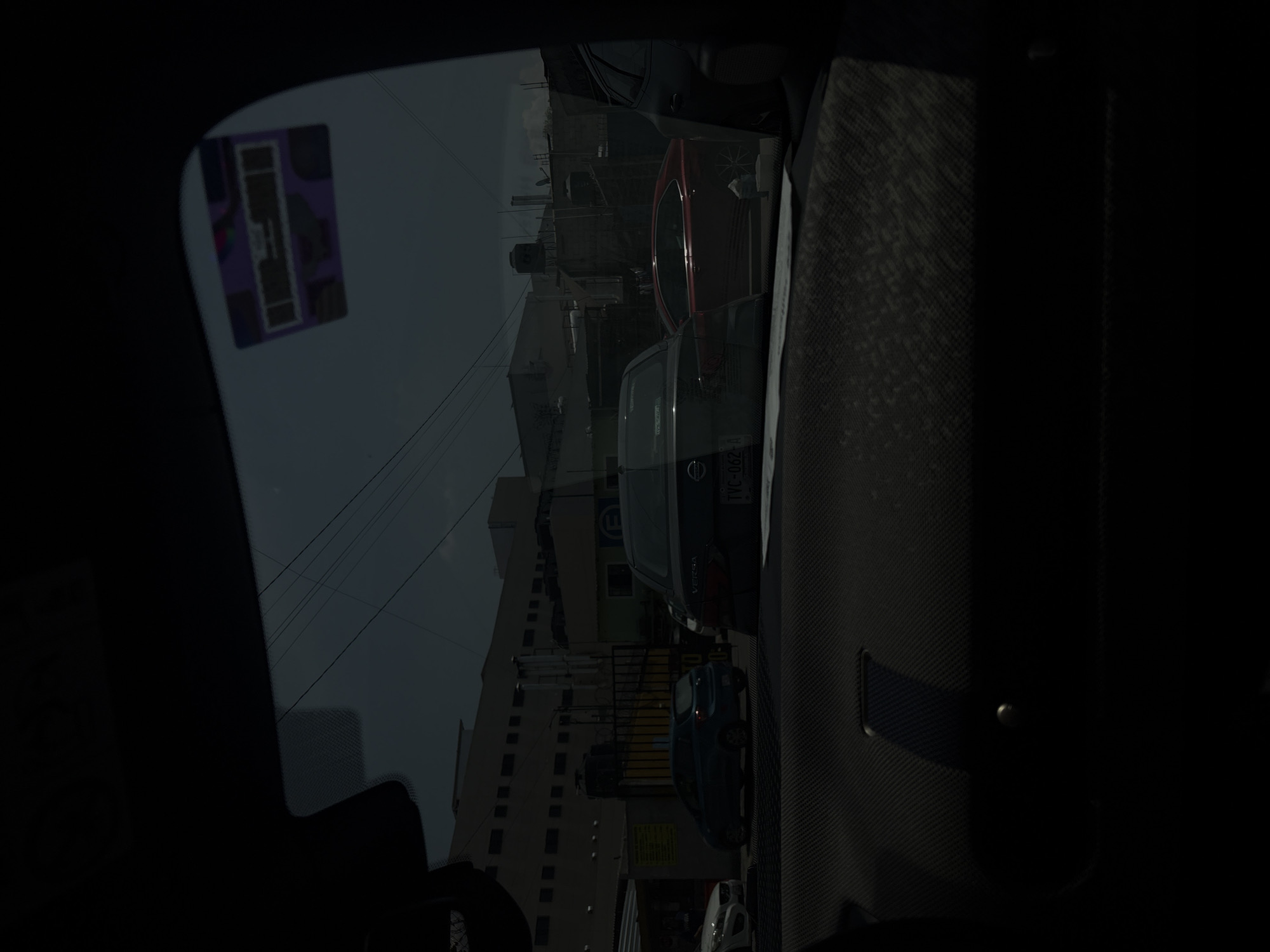 The image, taken from inside a car, shows a partial view of the vehicle's interior and the street outside. A sticker is visible on the windshield. The scene outside consists of a street with several parked cars, including a grey Nissan Versa with license plate "TVC-062-A" and a blue car. Buildings line the street, including a tall white building with dark windows and air conditioning units. Electrical wires span the street. The location is in Puebla, Mexico.