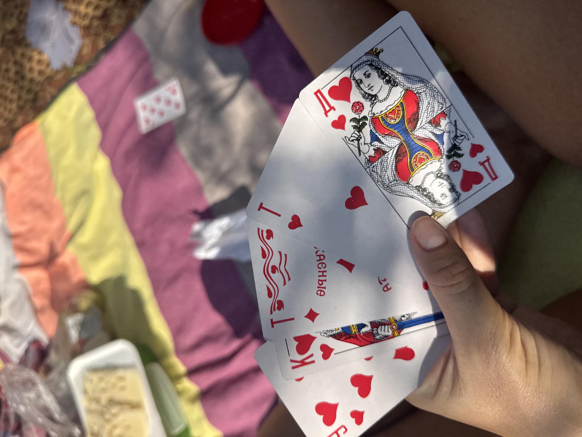 The image shows a close-up view of a person's hand holding several playing cards. The cards are of the suit of hearts and include the Queen of Hearts, an Ace of Hearts, and possibly other cards partially obscured. The Queen of Hearts card prominently displays an illustration of a queen holding a rose. The cards are fanned out, with the hand gripping them from the bottom.
In the background, a striped blanket or cloth is visible, featuring bands of pink, yellow, and orange. A white plastic container with some sort of food item, possibly cheese, is placed on the blanket. A red object, possibly a ball or a piece of fruit, is also visible in the upper left corner. The overall scene suggests an outdoor setting, possibly a picnic or a casual gathering on a sunny day, indicated by the bright lighting and shadows. The presence of playing cards suggests a game or leisure activity. No specific text is clearly legible on the playing cards or other objects in the image, except for what appears to be Cyrillic lettering on one of the cards in the background, possibly "КАСНЫЕ" or similar, which is common on Russian playing cards. The context of Moscow, Russia, is provided.