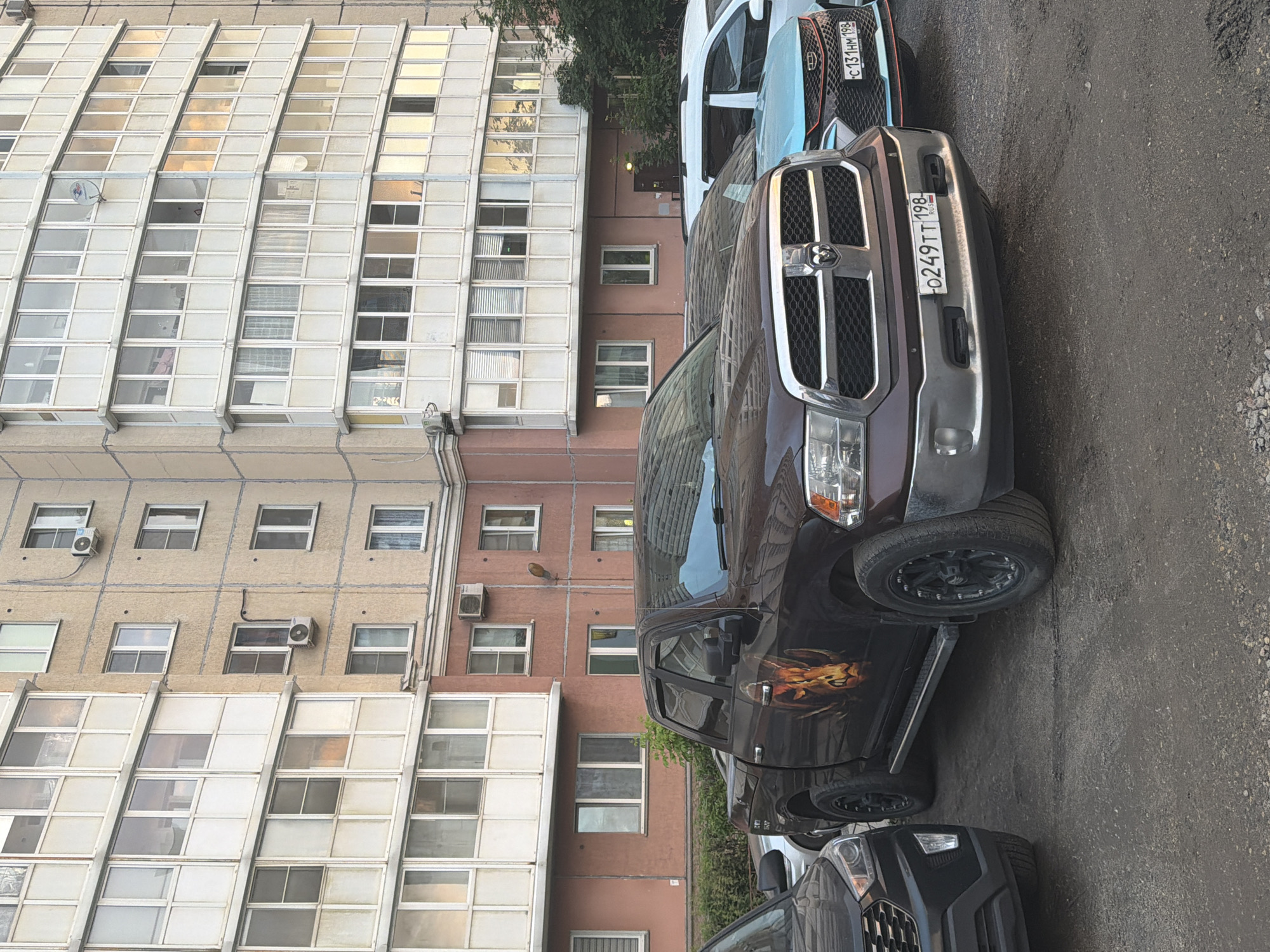 In a paved urban area, a dark brown Ram pickup truck with a graphic of a ram's head on its side is parked in front of a multi-story apartment building. The building features rows of windows and balconies, some with opaque coverings and others showing interior lights. To the left of the pickup, the front of a black SUV is visible. To the right of the pickup, a light blue compact SUV is parked, with its license plate starting with "C 131 HM 198". The pickup truck's license plate is "O249TT 198 RUSSIA". The ground is a mixture of dirt and gravel. The lighting suggests it is late afternoon or early evening.