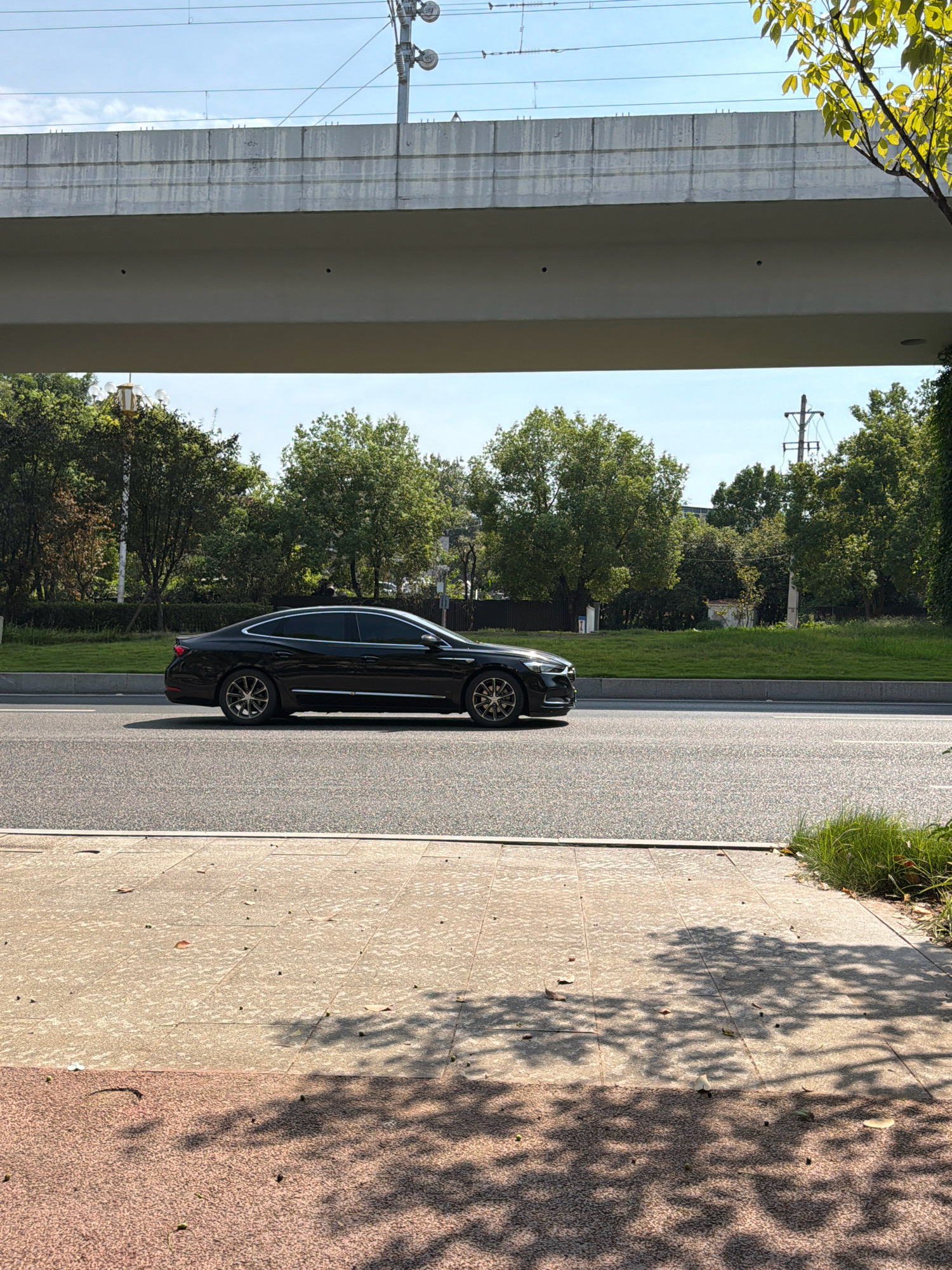 A black sedan is captured in profile, traveling rightward on a paved road beneath an elevated concrete structure. The vehicle, with tinted windows and chrome accents, showcases a sleek, modern design. Beyond the road, a lush green median separates it from a grassy embankment lined with mature trees. In the background, utility poles and electrical wires are visible against a bright blue sky with scattered clouds. The foreground features a pedestrian walkway constructed of light-colored square tiles and a section of darker, textured pavement, both dappled with the shadows of overhead foliage. The scene appears to be located in Jiangjin, China, suggested by its urban infrastructure and abundant greenery.