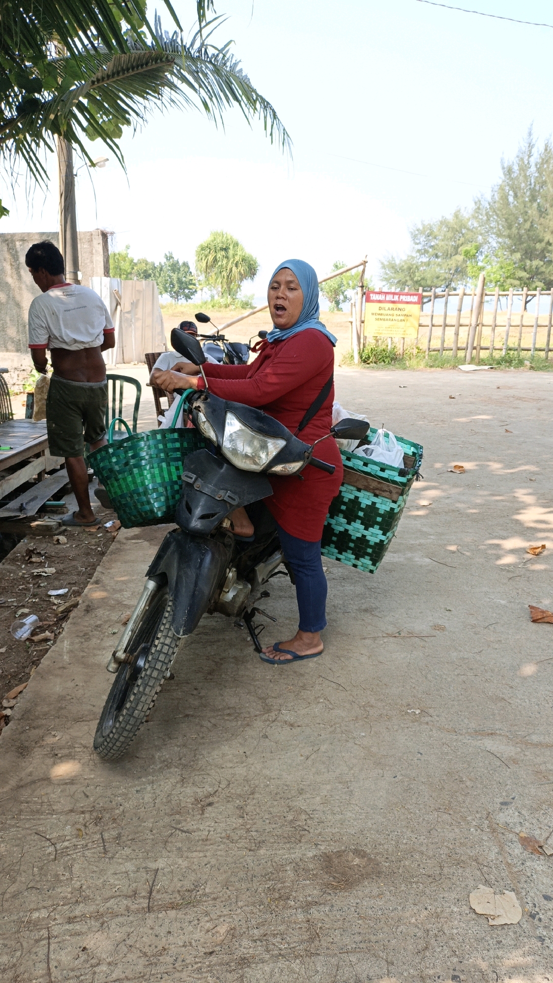 A full shot captures a woman in a red long-sleeved shirt and blue hijab, seated on a black motorcycle in Cikal, Indonesia. She appears to be speaking with an open mouth, her right hand resting on the handlebar. The motorcycle is equipped with two green woven baskets, one in front holding white plastic bags and another behind the seat. She wears blue jeans and blue flip-flops. To the left, a man in a white t-shirt and green shorts stands near a wooden structure, looking away from the camera. In the background, a yellow sign is posted on a wooden fence, indicating "Tanah Milik Pribadi Dilarang Membuang Sampah Sembarangan." The environment is outdoors, with a paved ground, some fallen leaves, and a palm tree to the left. The sky is clear and bright.