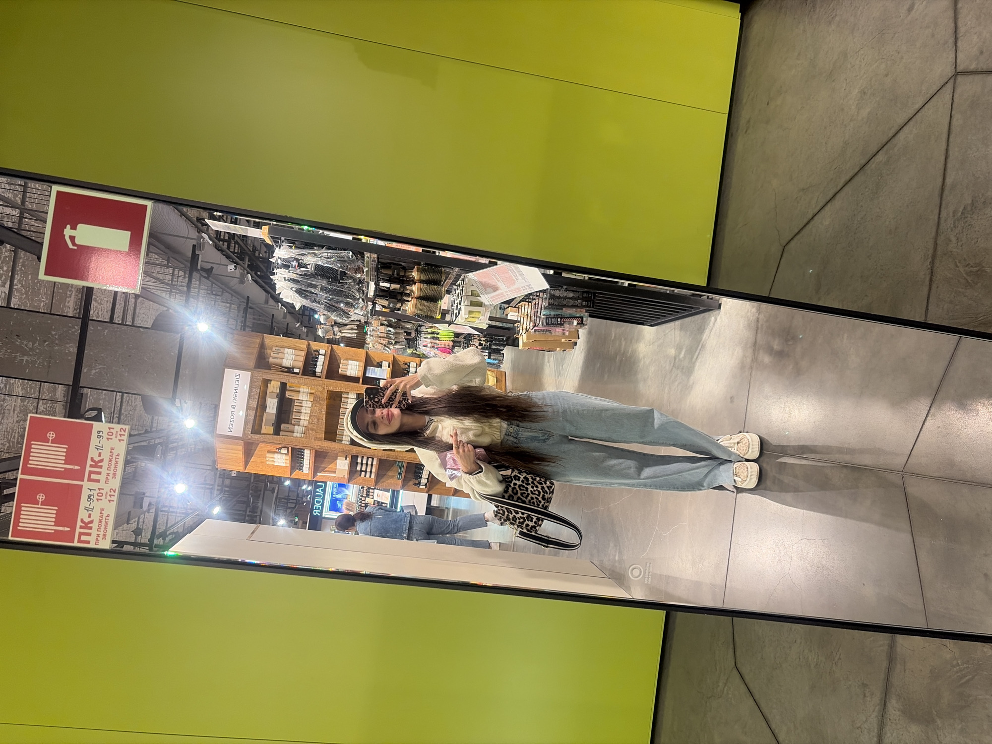 In the city of Saint Petersburg, Russia, a young woman is reflected in a tall, full-length mirror, taking a selfie. She is wearing a cream-colored hooded jacket, light blue wide-leg jeans, and patterned sneakers. She is holding a leopard-print handbag with a black strap and a small pink package in her left hand. Her right hand is holding a smartphone, angled to capture the reflection. The background visible in the mirror shows a retail environment, with shelves stocked with various products, possibly cosmetics or personal care items. Two red signs are visible hanging from the ceiling, one displaying fire extinguisher symbols and the other fire hose symbols with text in Russian. The walls of the store are painted a vibrant lime green. The floor is made of concrete tiles.