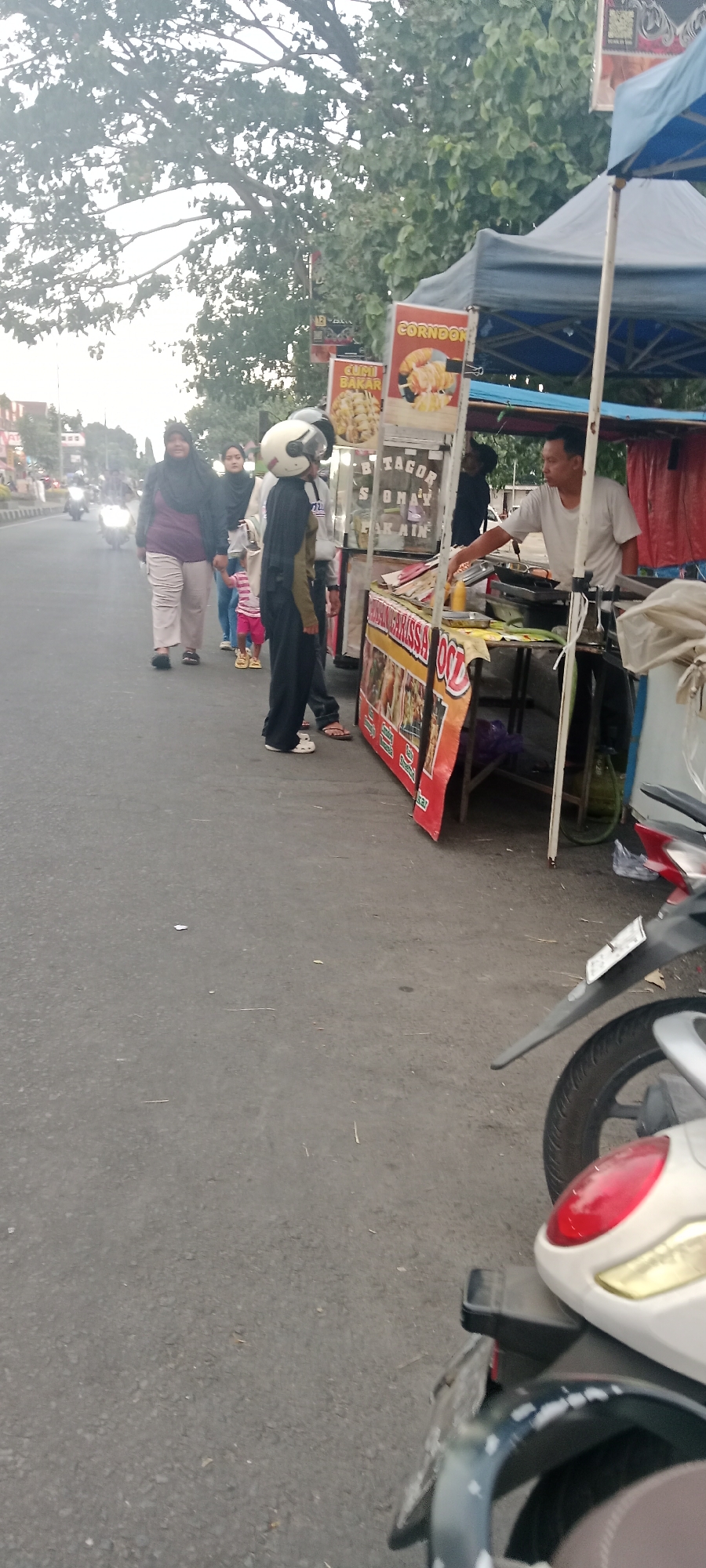 The image shows a street scene in Ngemplak, Indonesia, with a food stall on the right side of the frame. A man in a white t-shirt is behind the counter, appearing to be serving a customer or preparing food. The food stall has several signs with Indonesian text, including "CORNDON", "CUMI BAKAR", and "TACO". There are various food items visible on the counter, along with cooking equipment. In front of the stall, several people are walking or standing. A person wearing a dark robe and a white helmet is in the foreground, facing away from the camera. Behind them, a woman in a purple top and hijab, and another person in a blue shirt and pink striped shorts are walking. A motorcycle is parked on the right side of the image, partially obscuring the food stall. The street appears to be paved, and trees with green foliage are visible in the background. The overall lighting suggests it is daytime.