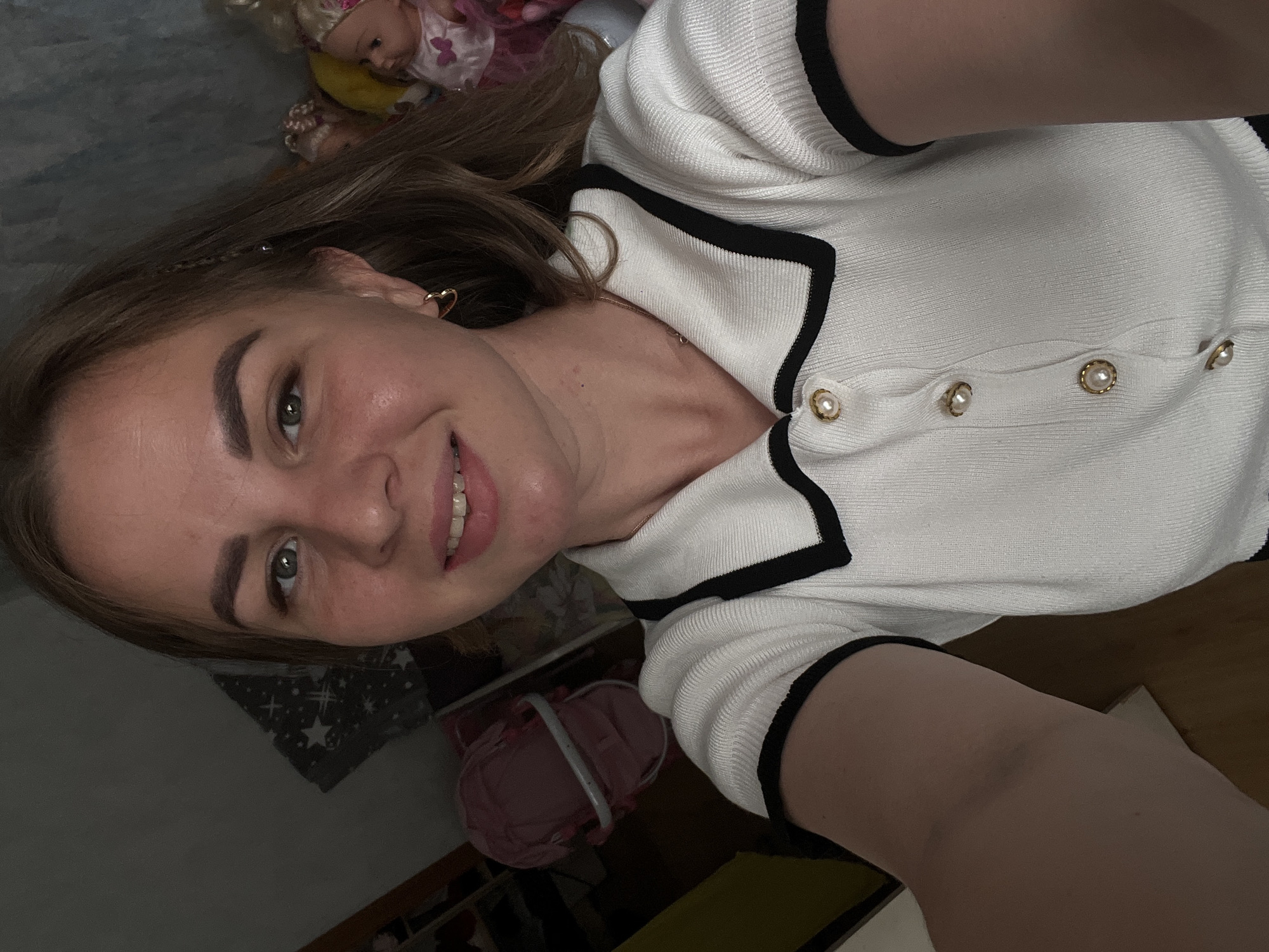 A selfie shows a woman with brown hair and green eyes smiling at the camera. She is wearing a white, short-sleeved shirt with black trim and pearl buttons. In the background, there are dolls, a gray surface with white stars, and a pink object. The location is assumed to be Izhevsk, Russia.