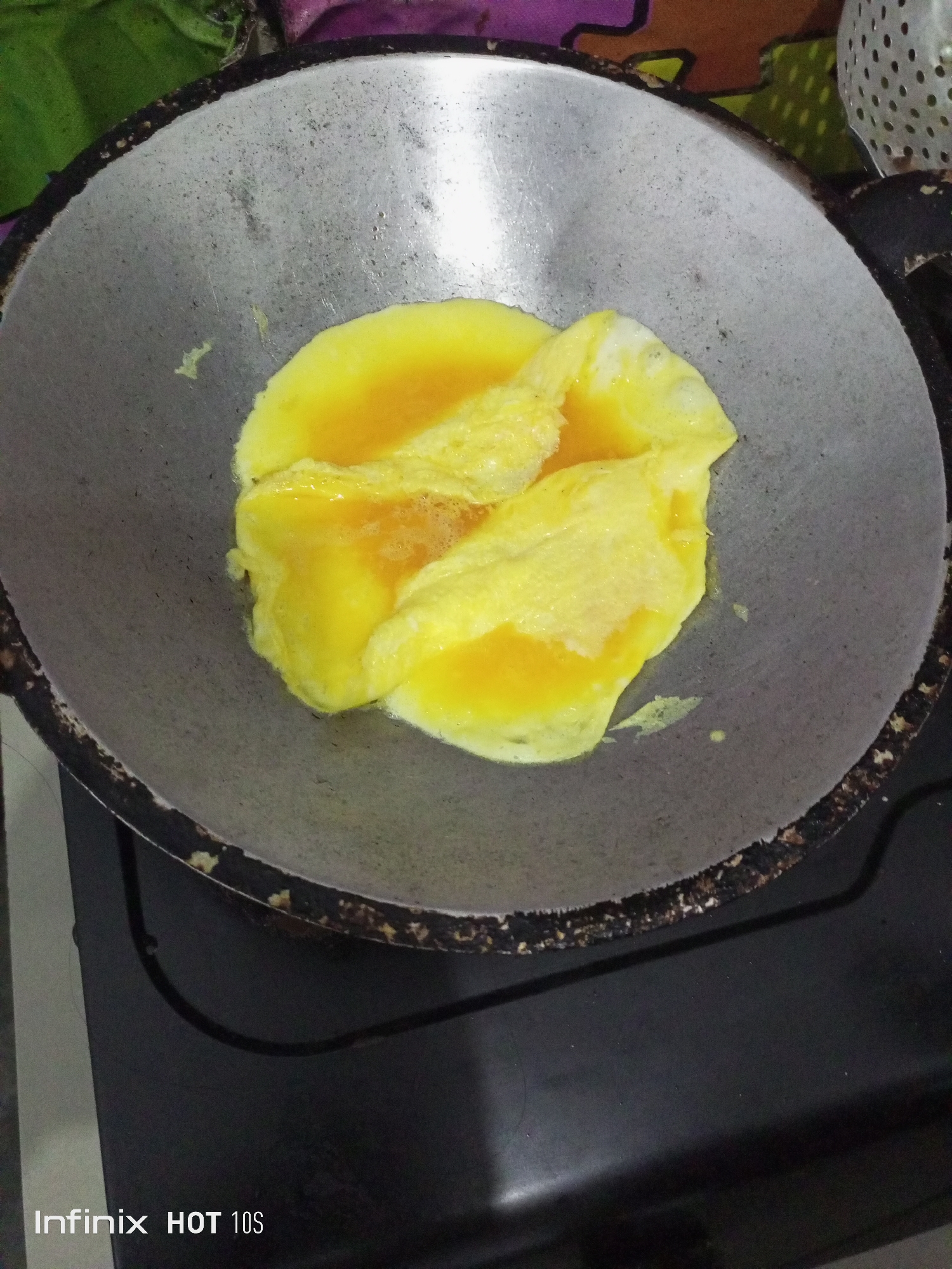 An overhead shot shows a round, well-worn metal frying pan on a black stove. Inside the pan, a bright yellow omelet is being cooked. The omelet appears to be folded over itself, and some of the liquid egg yolk is still visible. The frying pan has a dark, burnt rim. The background is out of focus, but suggests a kitchen environment with colorful patterns and a green leafy object. The bottom left corner of the image shows the text "Infinix HOT 10s".