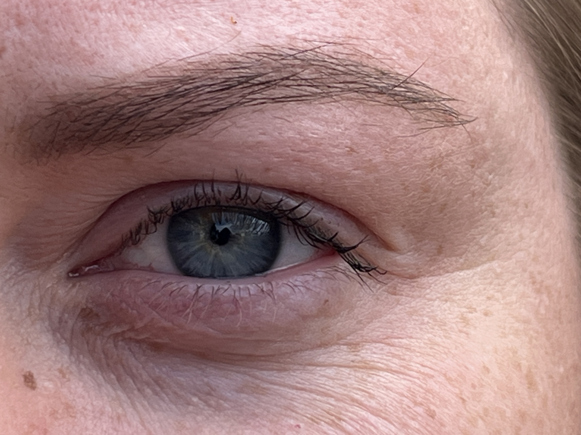 This image is a close-up of a human eye and eyebrow. The eye has blue iris and eyelashes with mascara. The eyebrow is dark and somewhat sparse. The skin around the eye shows wrinkles and blemishes. There is a mole visible on the skin below the eye. The location in the city of Ширяево, Russia is not specified, nor are any people, subjects, objects, actions, or interactions described in this context.