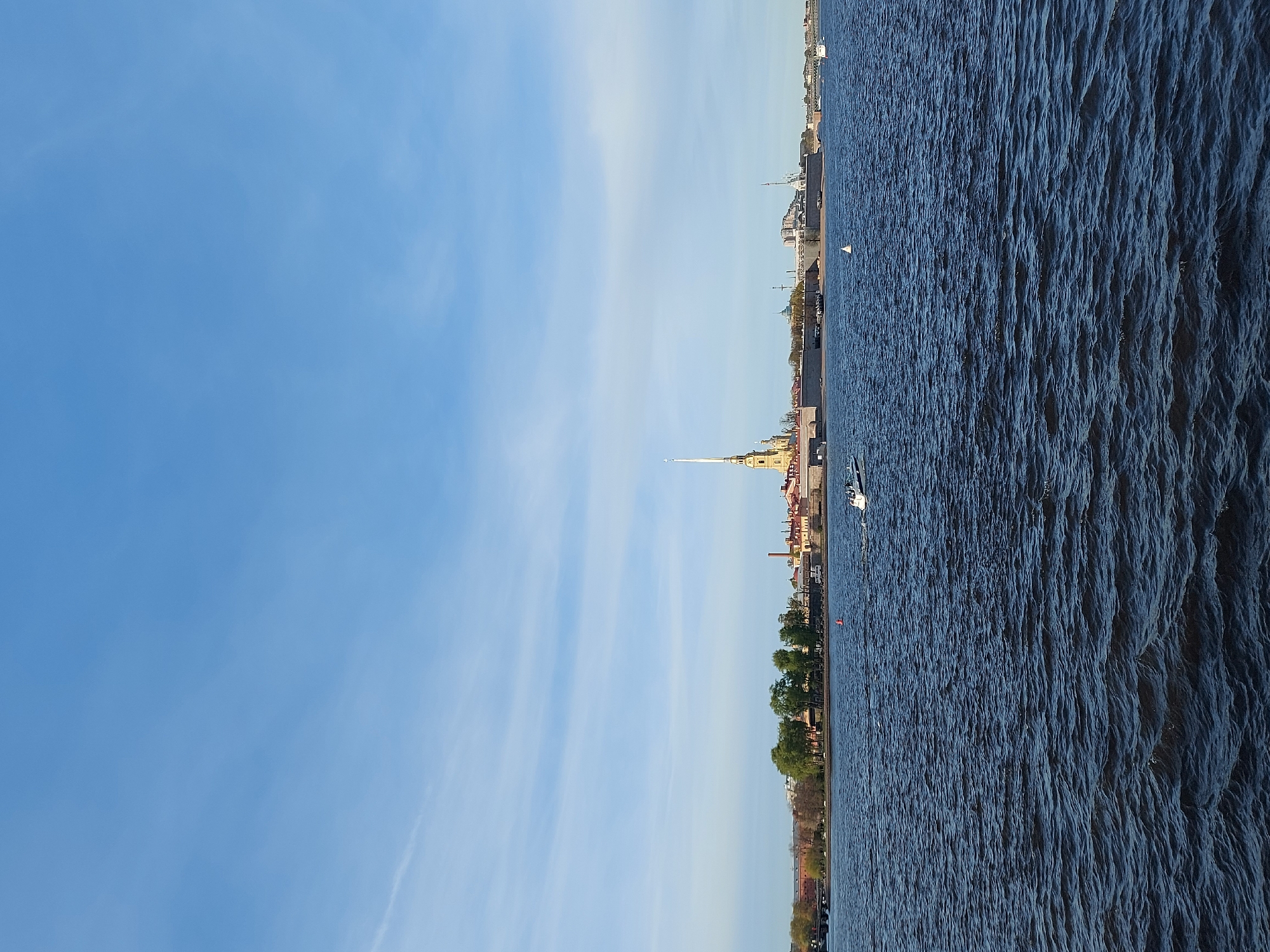 The image shows a waterfront view of Saint Petersburg, Russia. The sky is blue with thin clouds. The water of the Neva River is choppy. The cityscape includes trees, buildings, and the distinctive spire of the Peter and Paul Cathedral. A white boat is visible on the water. The image is oriented vertically.