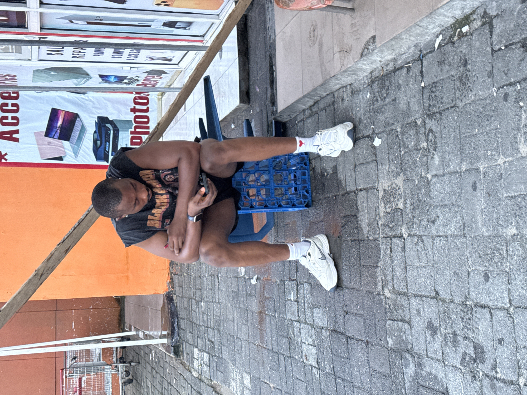 The image captures a man sitting on a blue plastic crate outside a shop in Ado, Nigeria. The man is wearing a black t-shirt, dark shorts, white socks with red hearts, and white sneakers. He is looking down at his phone, which he holds in his left hand. His arms are crossed. The shop behind him has an orange facade and displays signs advertising "Accessories," "SAL REPA," and "PHONE COMP ACCESS." There is a wooden plank leaning against the shop wall. In the background, to the left, are two red shopping carts. The ground is paved with gray bricks, and there are some wet patches, suggesting it may have recently rained. Debris, including crumpled paper and cigarette butts, is scattered on the ground.