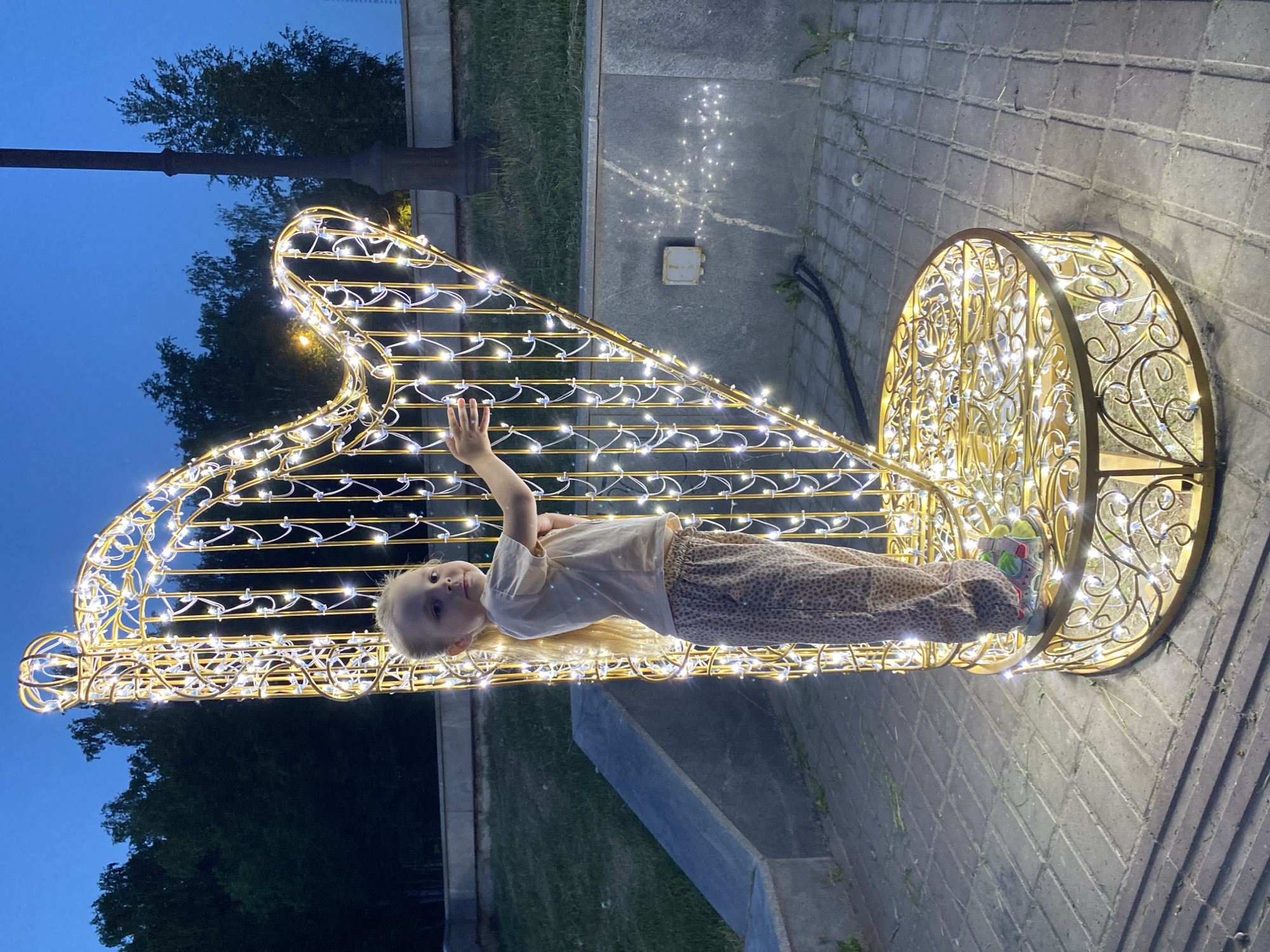 A young child, likely female with blond hair, lies horizontally across a large, golden harp sculpture illuminated with numerous small white lights. She wears a light-colored shirt and patterned pants. Her right arm is raised. The sculpture is located outdoors, seemingly in a public space at twilight. The setting is Votkinsk, Russia. The background includes a dark sky, trees, and a lamppost. The ground is paved with gray bricks.
