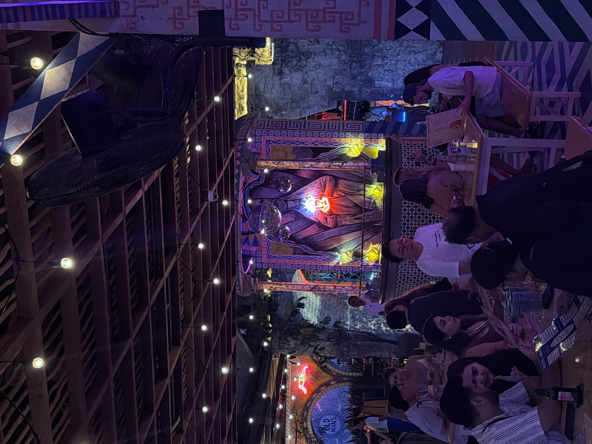 The image depicts the interior of a dimly lit restaurant or bar in North Kuta, Indonesia, with a vibrant, eclectic ambiance. The ceiling is constructed of exposed wooden beams, with numerous string lights creating a festive atmosphere. Dominating the background is a large, colorful mural or art installation featuring figures and geometric patterns, illuminated by neon and colored lights. Several people are seated at tables, engaged in conversation and looking at menus. The decor includes stone-like textured walls and decorative patterned panels. A prominent feature is a large, dark, possibly fan-like object hanging from the ceiling. The overall scene suggests a lively social gathering in a uniquely decorated establishment.