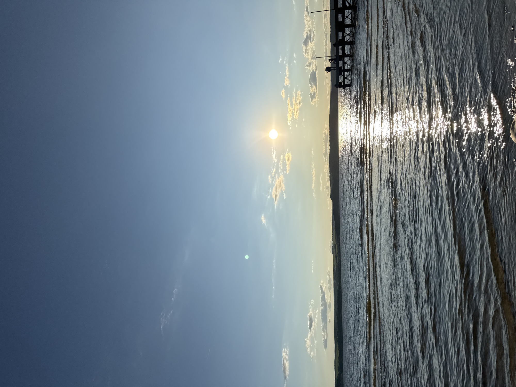 The image depicts a tranquil scene at a lake, possibly in Матюшино, Russia, during what appears to be late afternoon or early evening. The sun is low in the sky, casting a warm glow and creating a shimmering reflection on the water. A wooden pier extends into the lake on the right side of the frame, with two silhouetted figures visible on it, one standing and one seated. The water is calm, with gentle ripples reflecting the sunlight. In the background, a distant shoreline is visible, lined with trees and rolling hills under a clear blue sky dotted with a few fluffy white clouds. The overall atmosphere is peaceful and serene, evoking a sense of natural beauty and quiet relaxation.