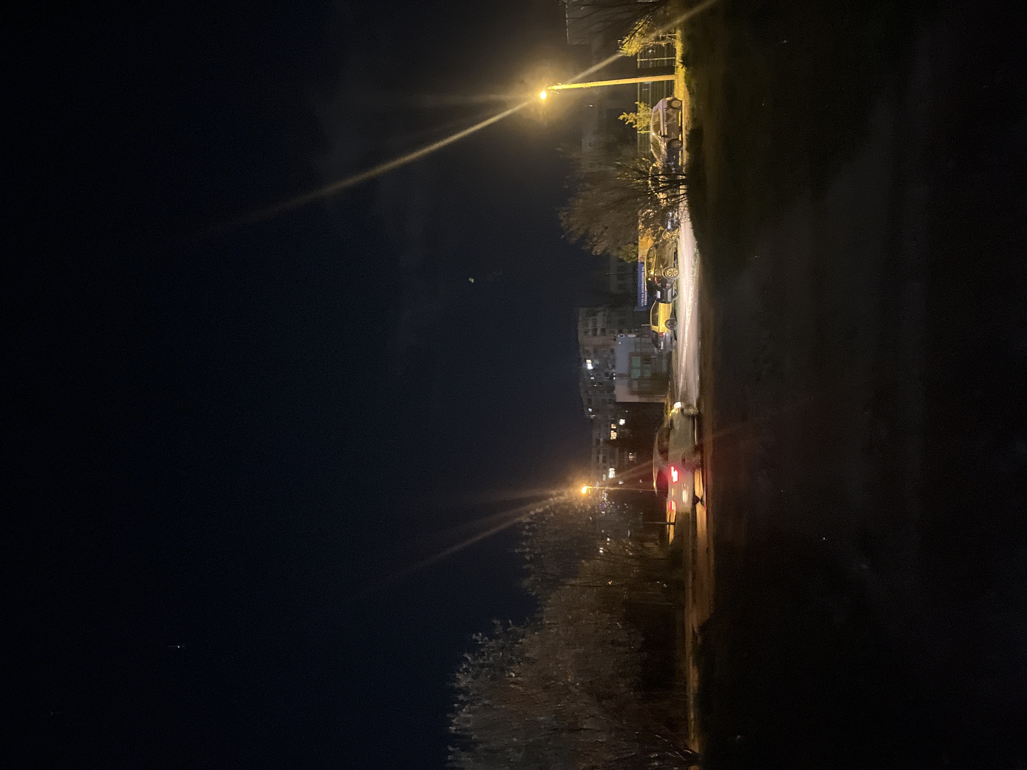 At night, a street scene shows a street light illuminating several parked cars and some buildings in the distance. One car has its brake lights on. The location appears to be in Sofia, Bulgaria.