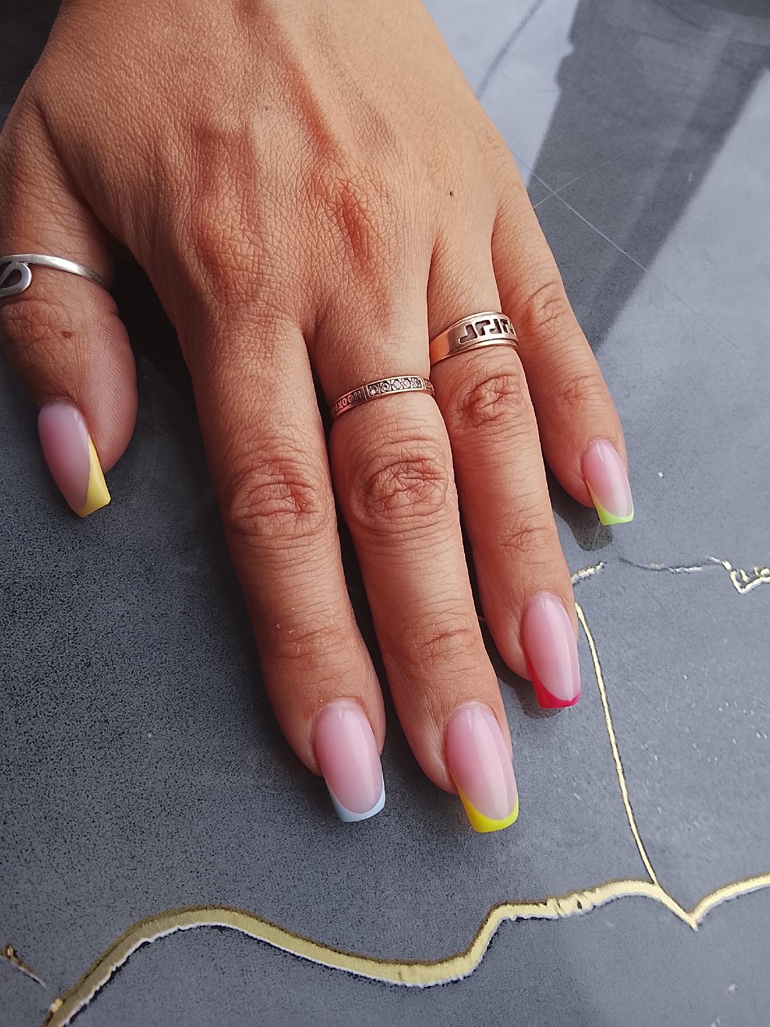The image displays a close-up view of a person's left hand adorned with colorful French-tip manicured nails. The nails feature a light pink base with vibrant tips in yellow, light blue, neon green, and red. Two rings are visible on the fingers: a thin band with small stones on the ring finger and a decorative gold-toned band with a Greek key pattern on the middle finger. A simple silver ring with a loop design is on the index finger. The hand rests on a dark gray textured surface with subtle gold linear accents. The image is a studio shot, likely taken in a nail salon. There is no identifiable information that would place this scene in Almaty, Kazakhstan.